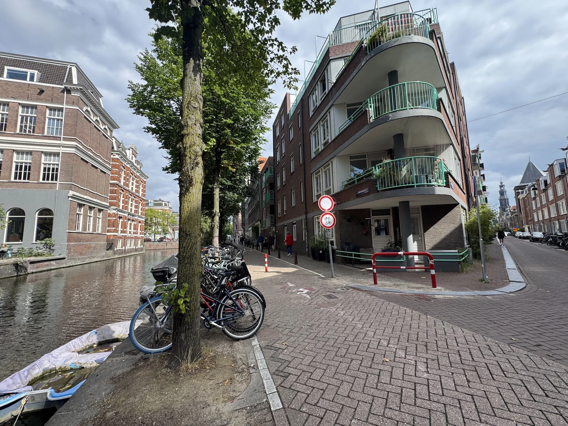 Bicycles are parked along the sidewalk next to a canal, with a large tree and a multi-story residential building with balconies nearby. The sky is partly cloudy, and there are a few pedestrians on the street.