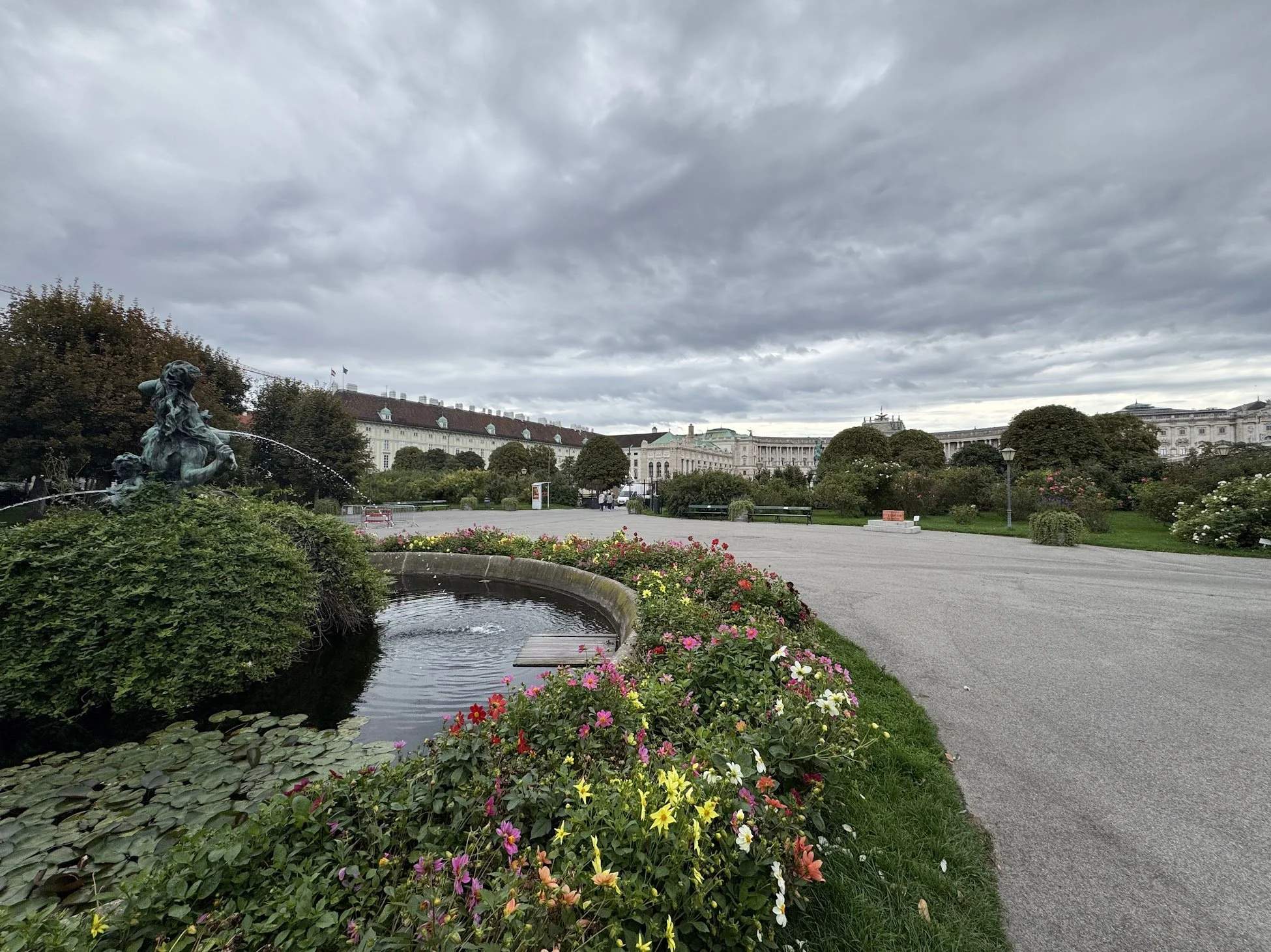 A city park with a fountain, colorful flowers, trees, historic buildings, and cloudy skies.