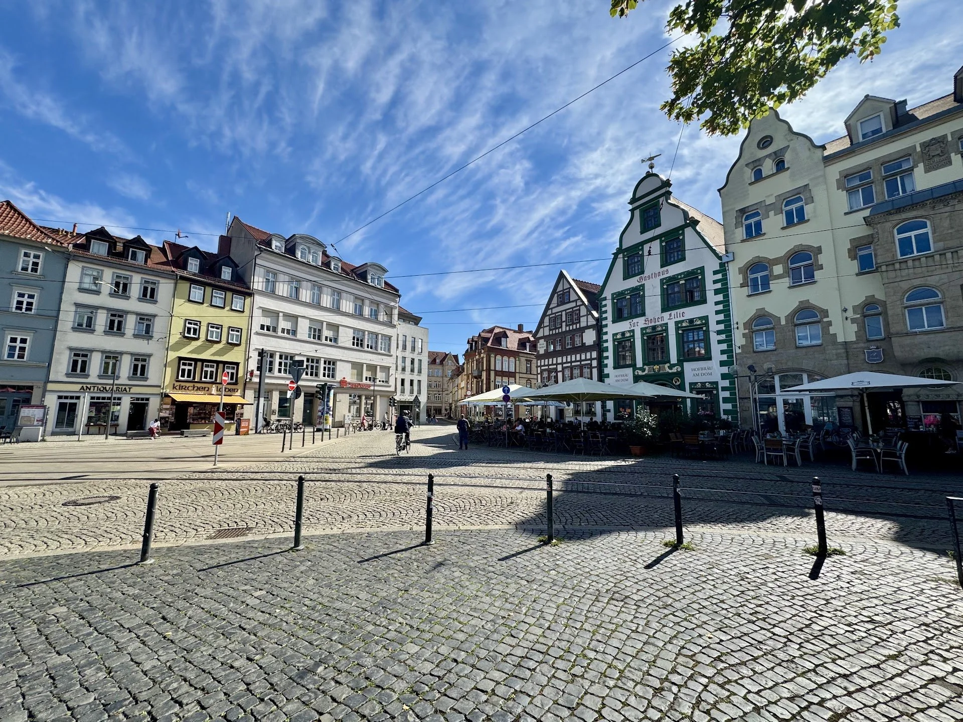 A European city square with cobblestone pavement, outdoor seating under umbrellas, and colorful historic buildings under a partly cloudy sky.