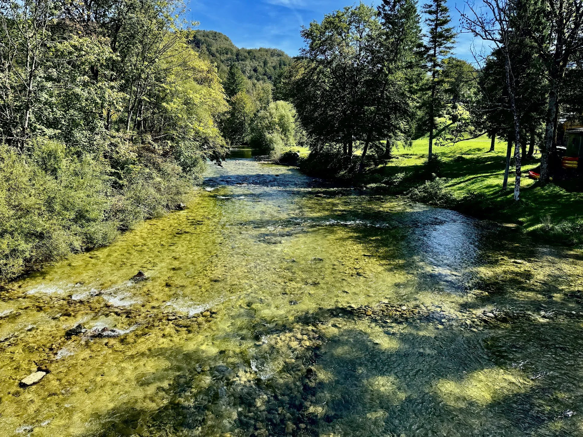 A scenic view of a clear river flowing through a forested landscape with trees on both sides, mountains in the distance, and a blue sky overhead.
