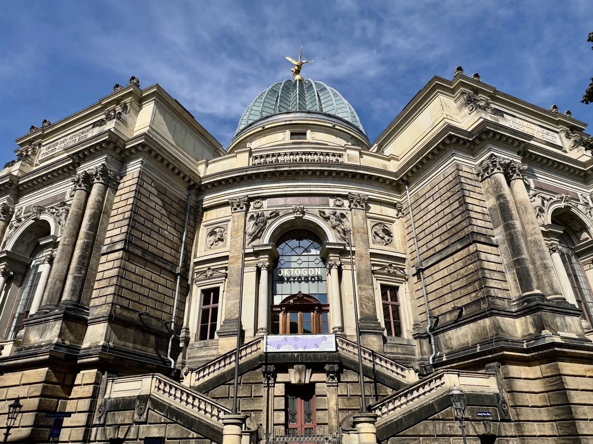 The facade of the historic Oktogon building with ornate architectural details, columns, and a glass dome topped with a golden statue against a blue sky.