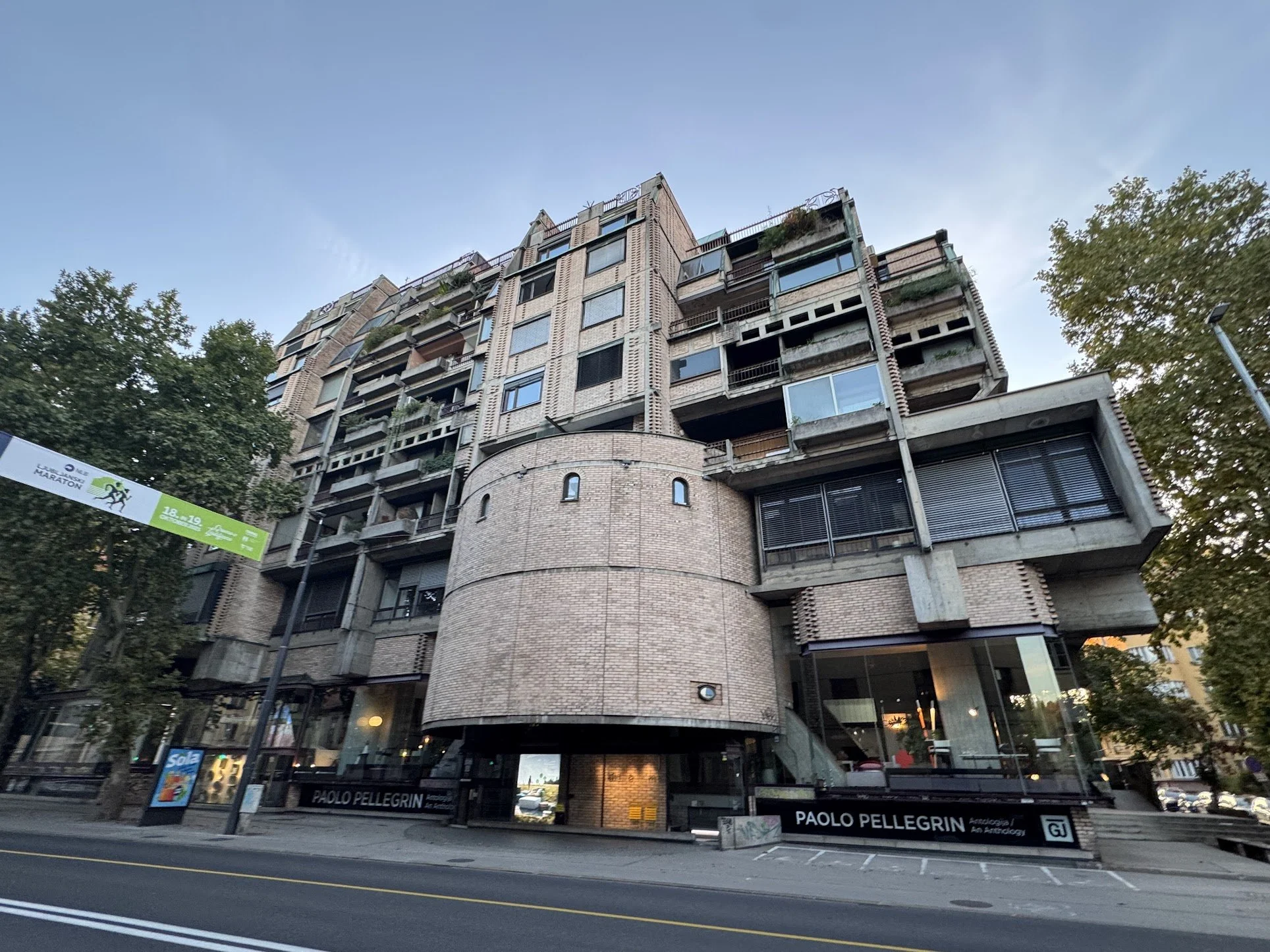 A tall multi-story building with a modern architectural design featuring brickwork, large windows, and balconies. There are trees in the foreground and a banner on the street level advertising a marathon event.