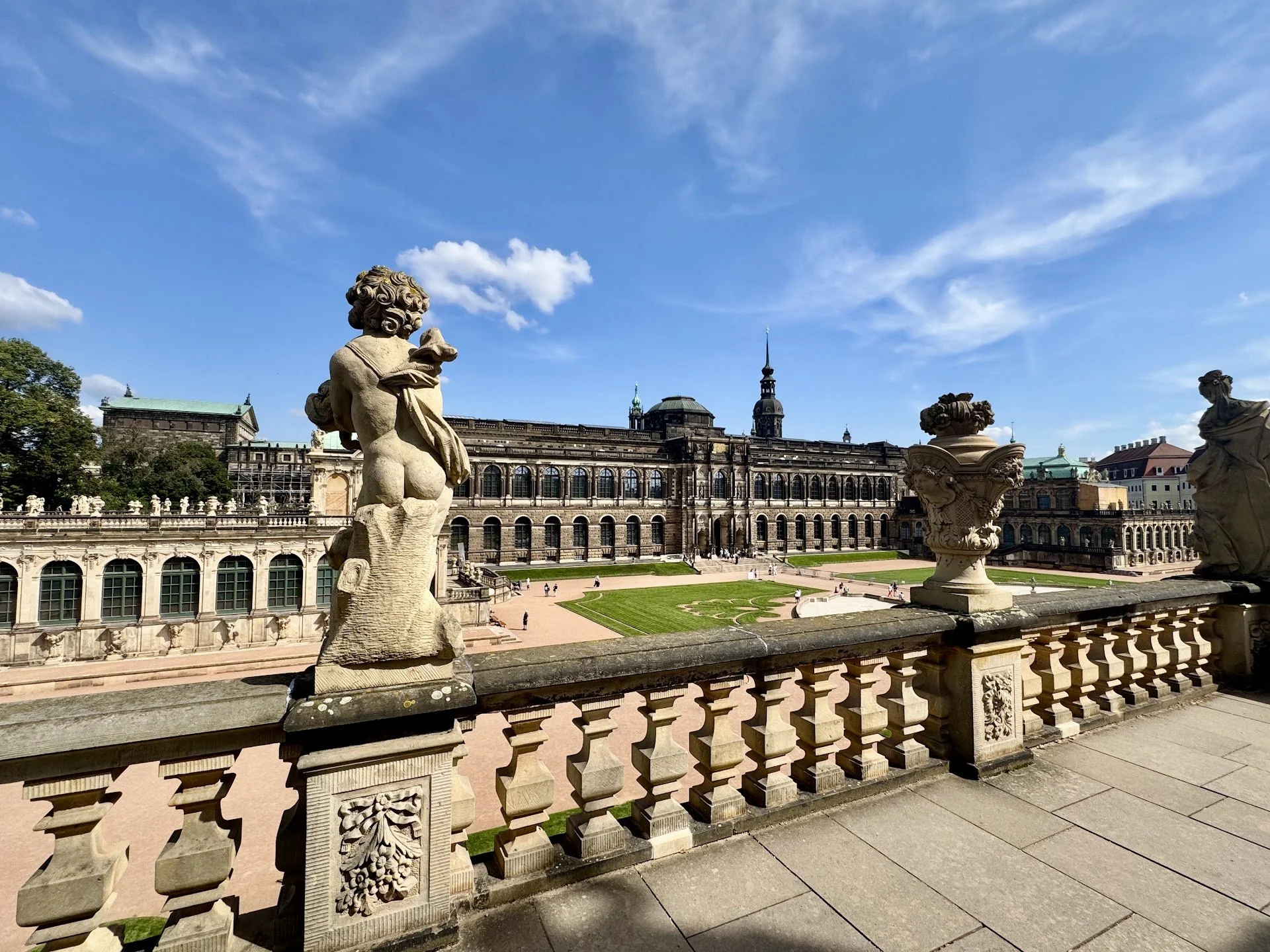 View of a historic palace with ornate stone balustrades and sculptures in the foreground, a large glass-covered courtyard, classical architecture, and a blue sky with some clouds.