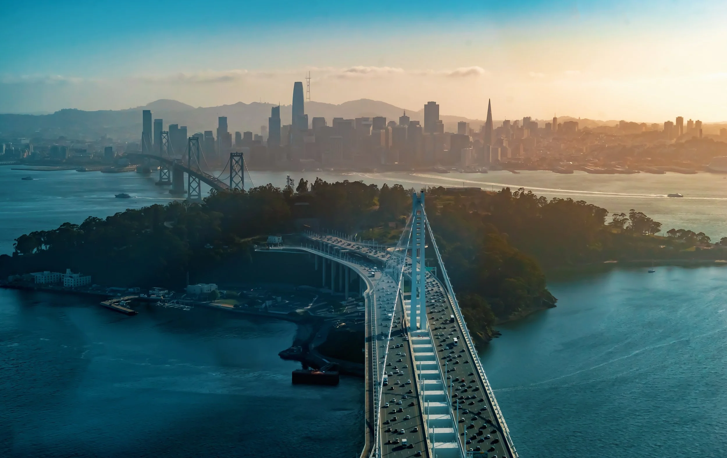 An aerial view of the San Francisco skyline with the Bay Bridge in the foreground, taken at sunset.