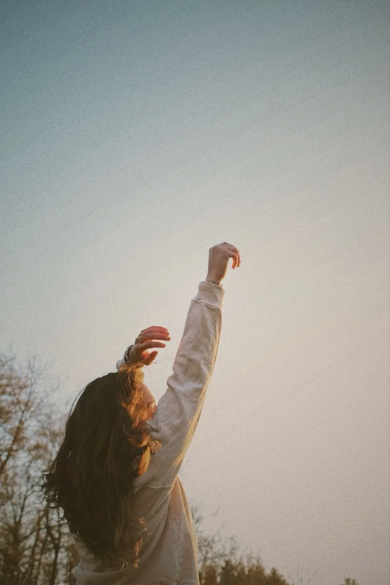 A person with long hair reaching up with one arm towards the sky during sunset or sunrise, with trees in the background.