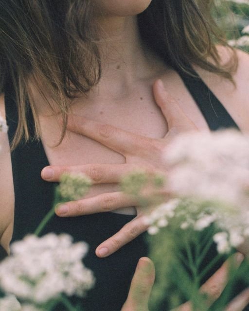 A person wearing a black sleeveless top holds their hand against their chest, with their fingers crossed. Flowers in the foreground slightly obscure the view.