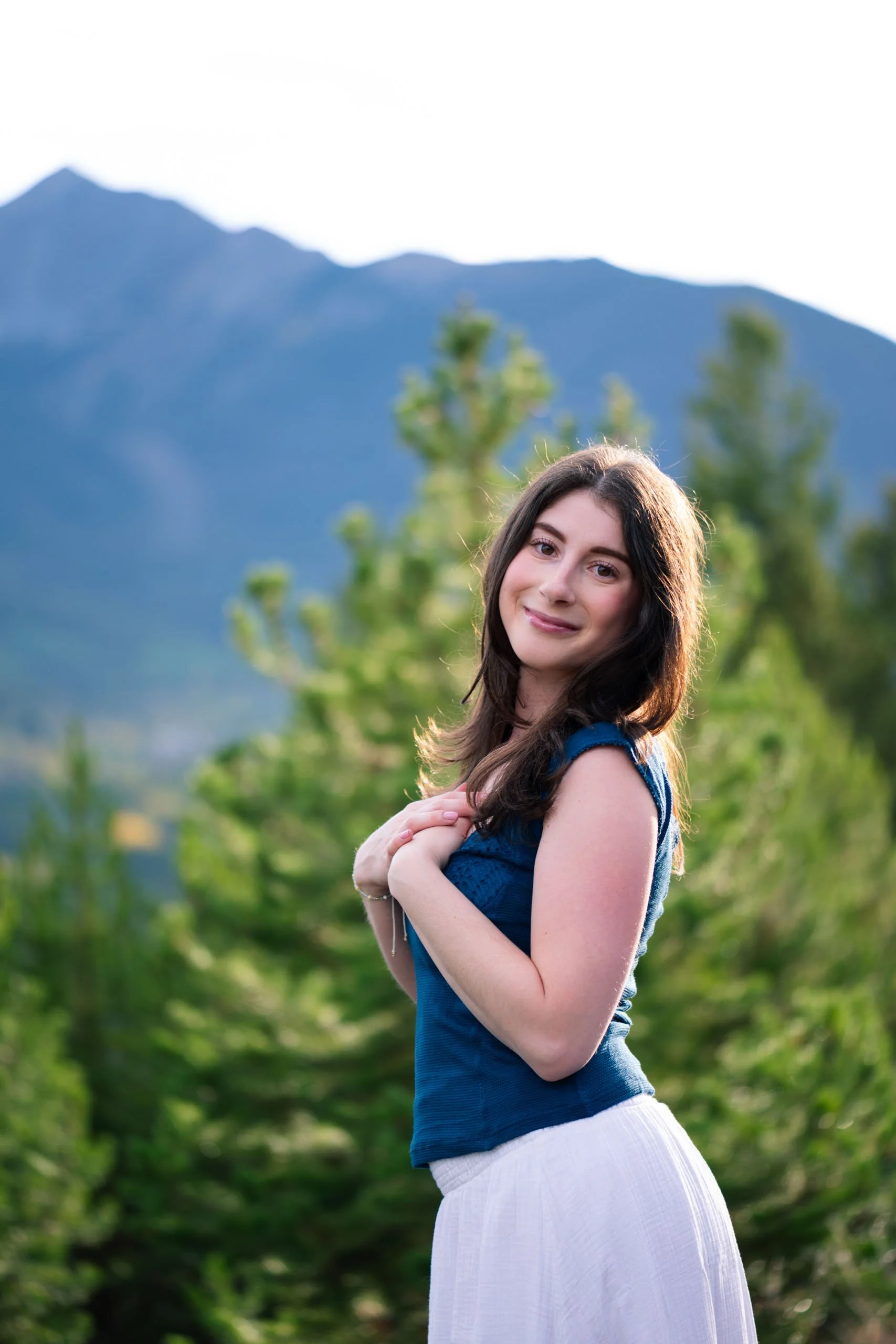 A young woman with long dark hair, smiling, standing outdoors with green trees and mountains in the background, wearing a blue top and a white skirt.