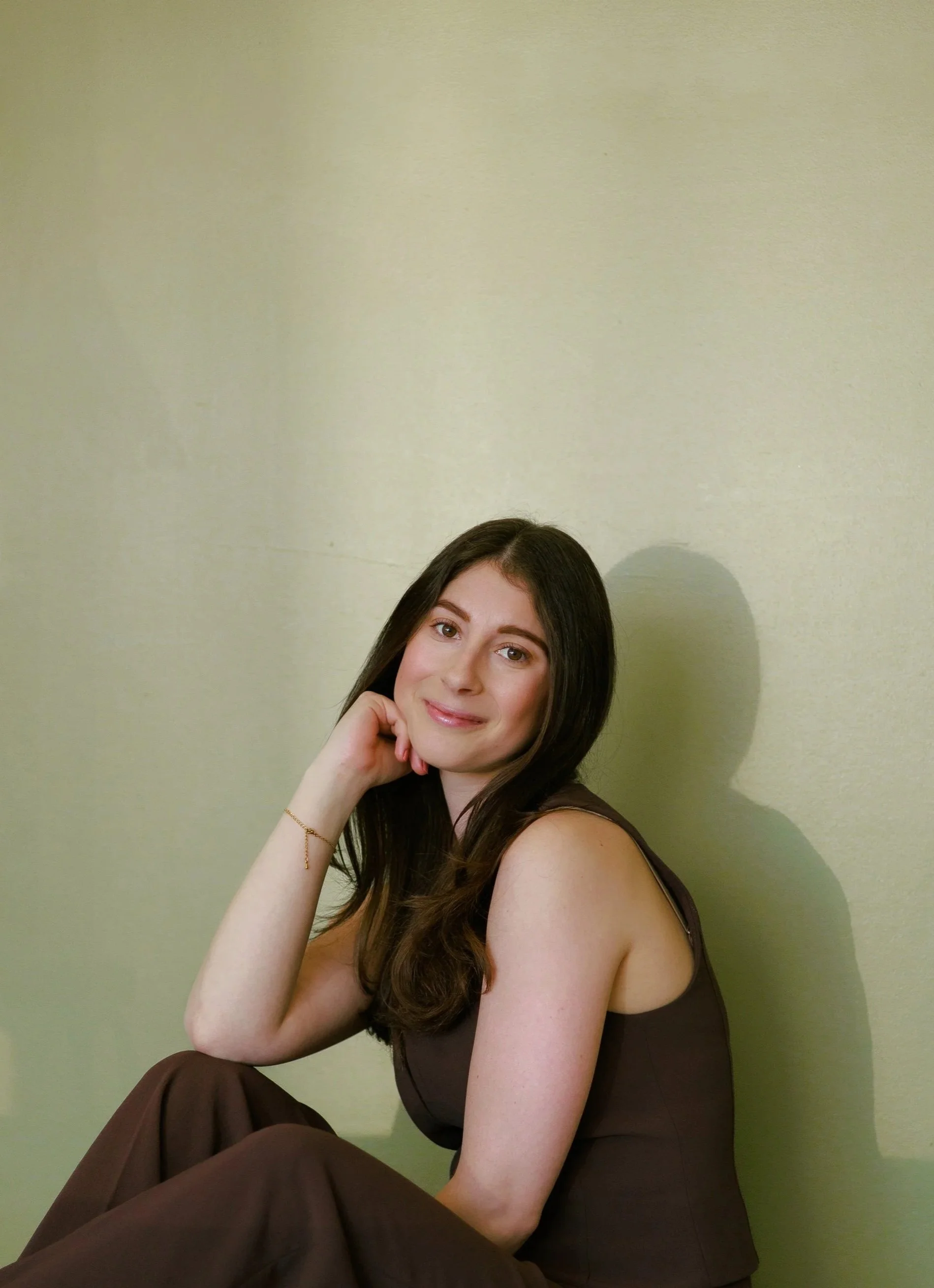 A woman with long brown hair, resting her chin on her hand, sitting against a pale green wall, wearing a sleeveless brown dress.