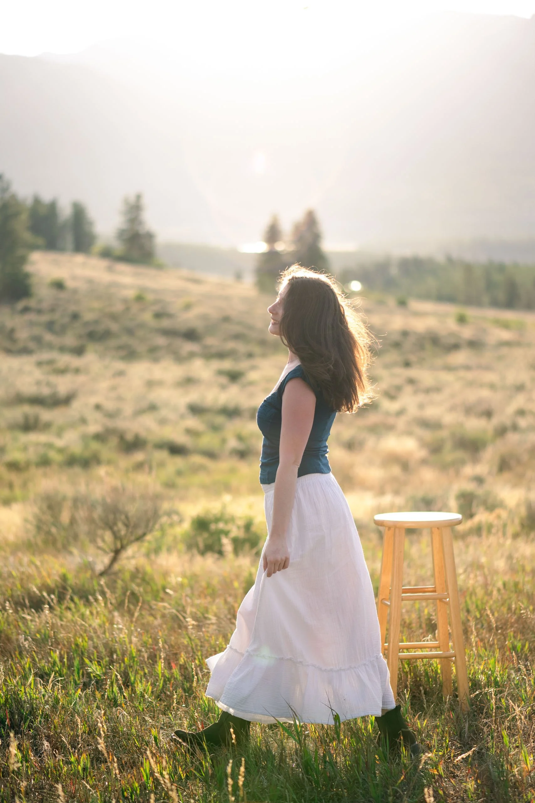 A woman with long brown hair in a braid, wearing a dark sleeveless top and a long white skirt, stands in a grassy field with mountains and trees in the background. The sunlight is bright, creating a warm glow, and there is a wooden stool on her right.