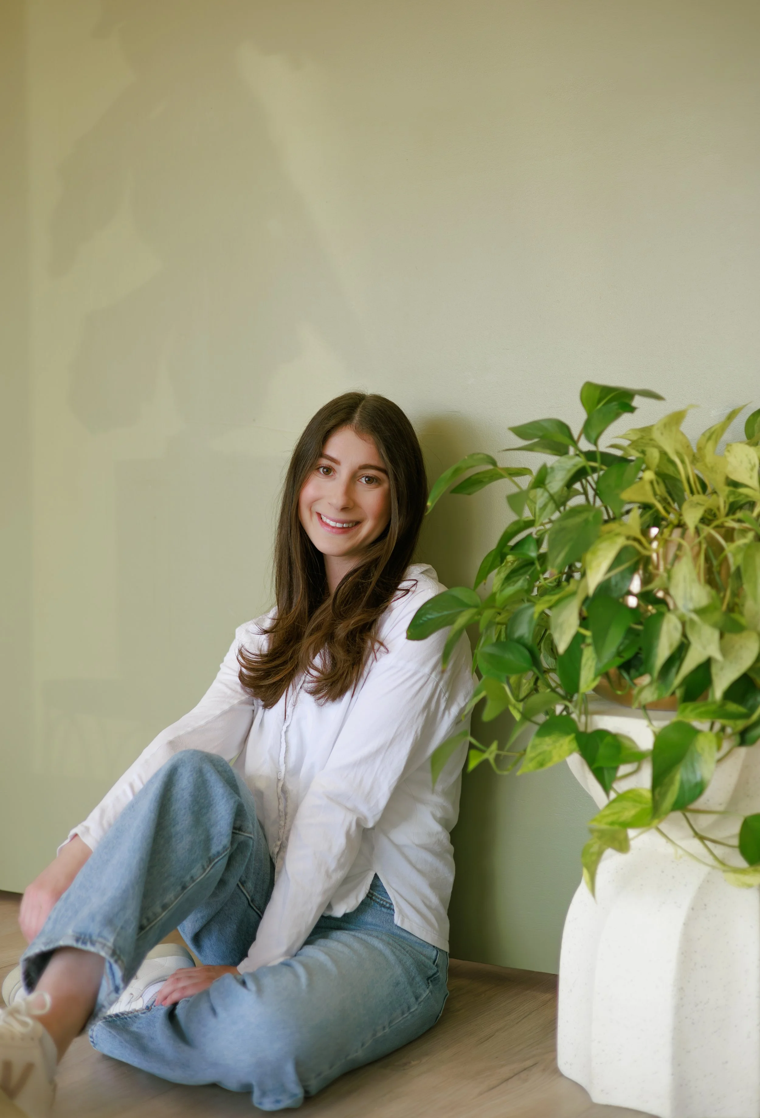 Smiling young woman with long brown hair sitting on the floor next to a large white pot with green variegated leaves, leaning against a light green wall.