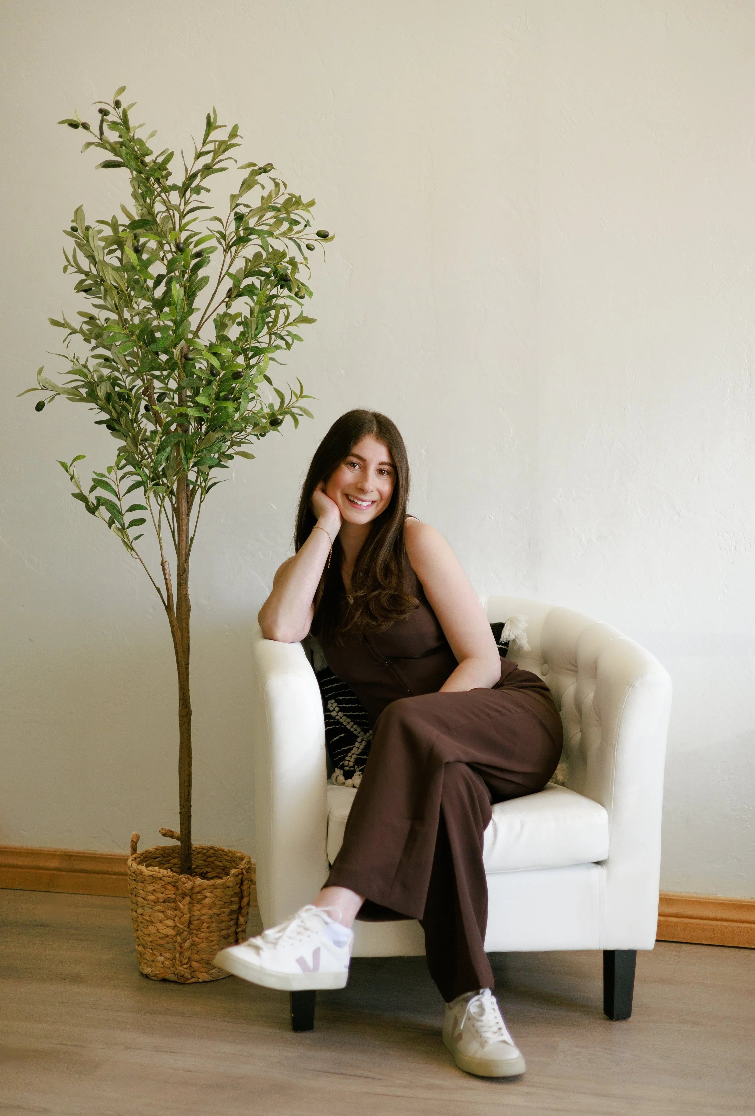 A woman with long dark hair sitting on a white armchair in a room with a light-colored wall, next to a tall potted plant with green leaves and small black berries. She is smiling, wearing a brown sleeveless outfit and white sneakers.
