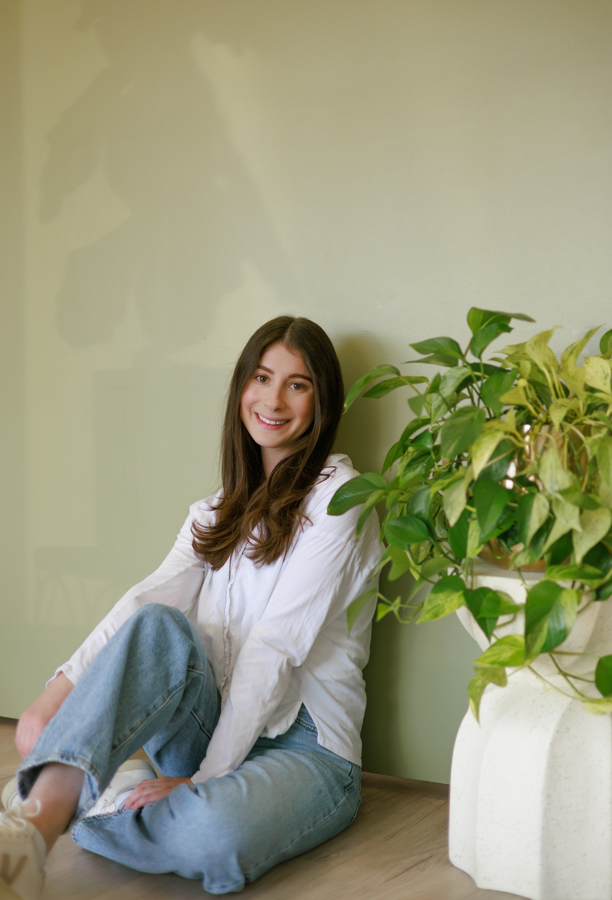 Young woman with long brown hair sitting on the floor next to a large potted green houseplant, smiling at the camera in a well-lit room with light-colored walls.