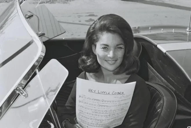 A woman with dark, wavy hair smiling and holding a handwritten letter inside a vintage car.