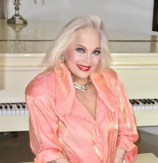 A woman with blonde hair and red lipstick sitting in front of a white piano, wearing a pink satin blouse with gold accents and a pearl necklace.