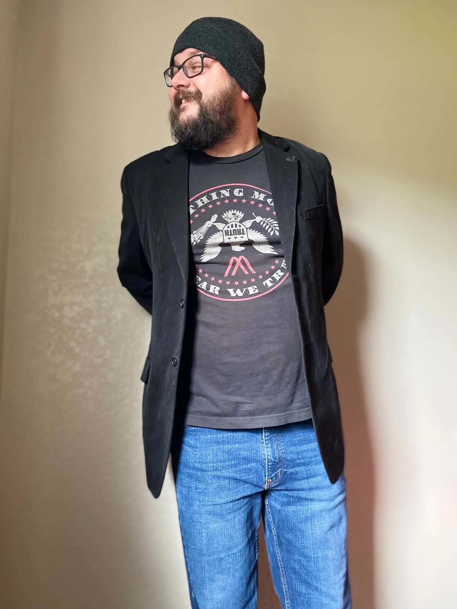 Rob Perez, Pink Crow Press co-founder, wearing a black knit beanie, glasses, a black blazer, a graphic t-shirt, and blue jeans, standing against a plain wall.