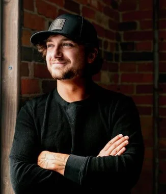 JD Justice, Pink Crow Press illustrator, with a beard and mustache wearing a black baseball cap and black shirt, standing with arms crossed in front of a brick wall, smiling and looking out a window.