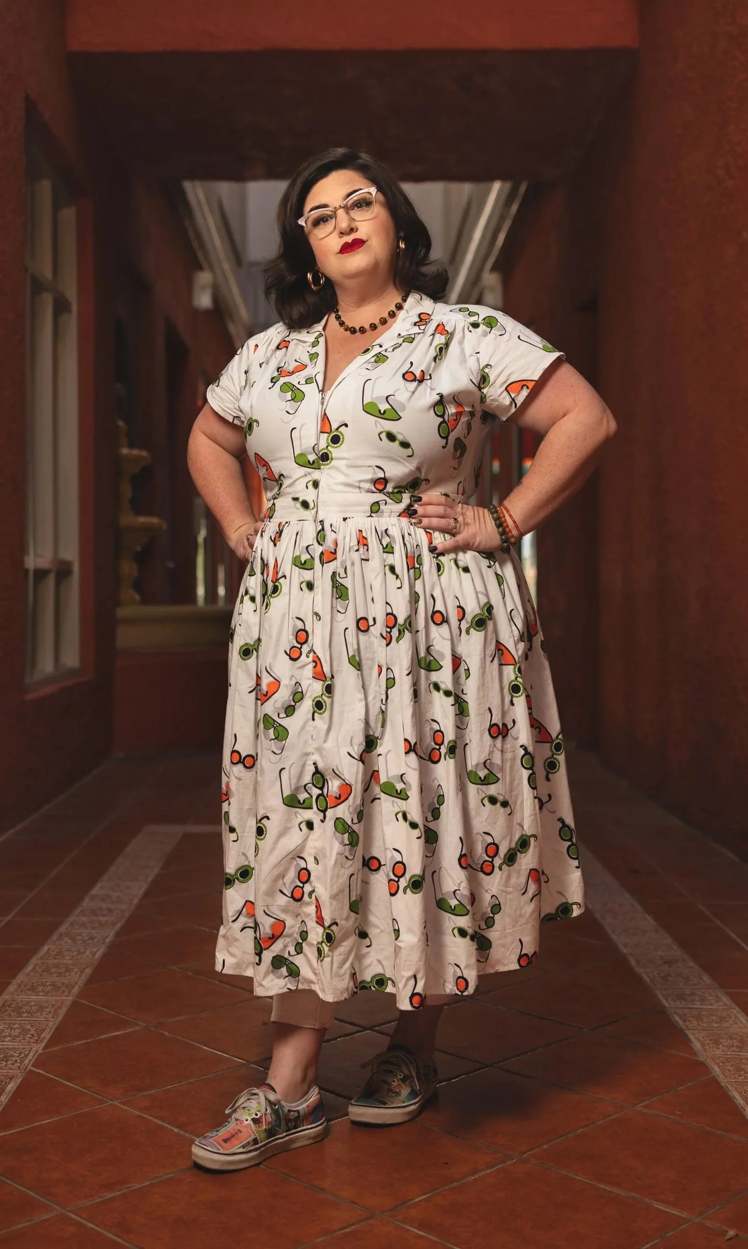 Pam Holmes, Pink Crow Press co-founder, with shoulder-length dark hair and glasses stands with hands on her hips in a hallway with brick floors and dark red walls.