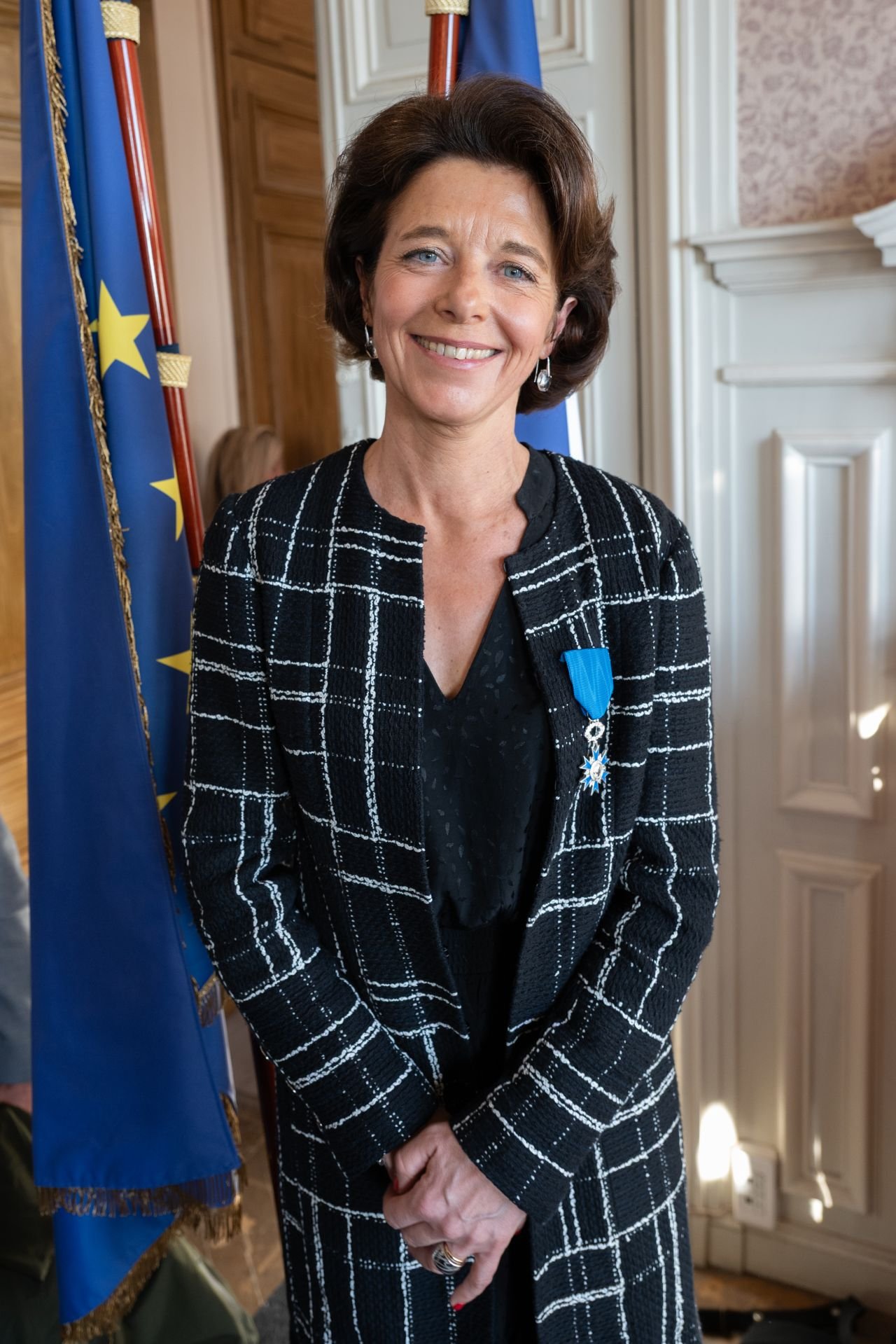 Une femme souriante portant une médaille de l'ordre de la Légion d'honneur, posant devant un drapeau européen dans une salle élégante.