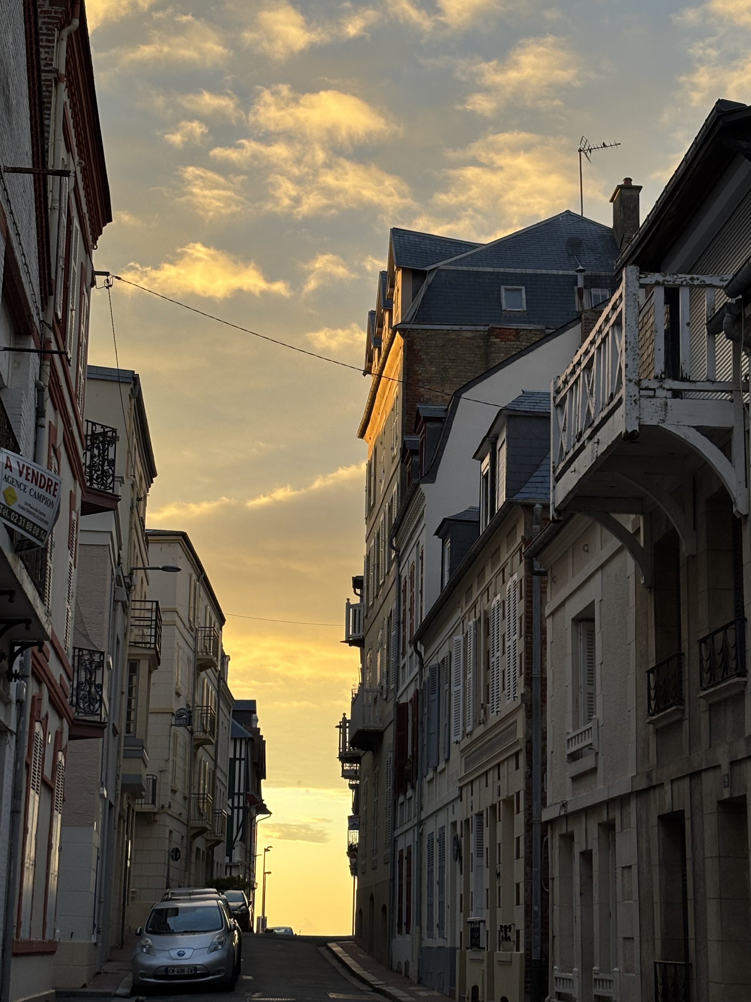 Rue pavée avec voitures stationnées, bâtiments résidentiels à plusieurs étages, balcons en fer forgé, ciel au coucher du soleil avec des nuages colorés.