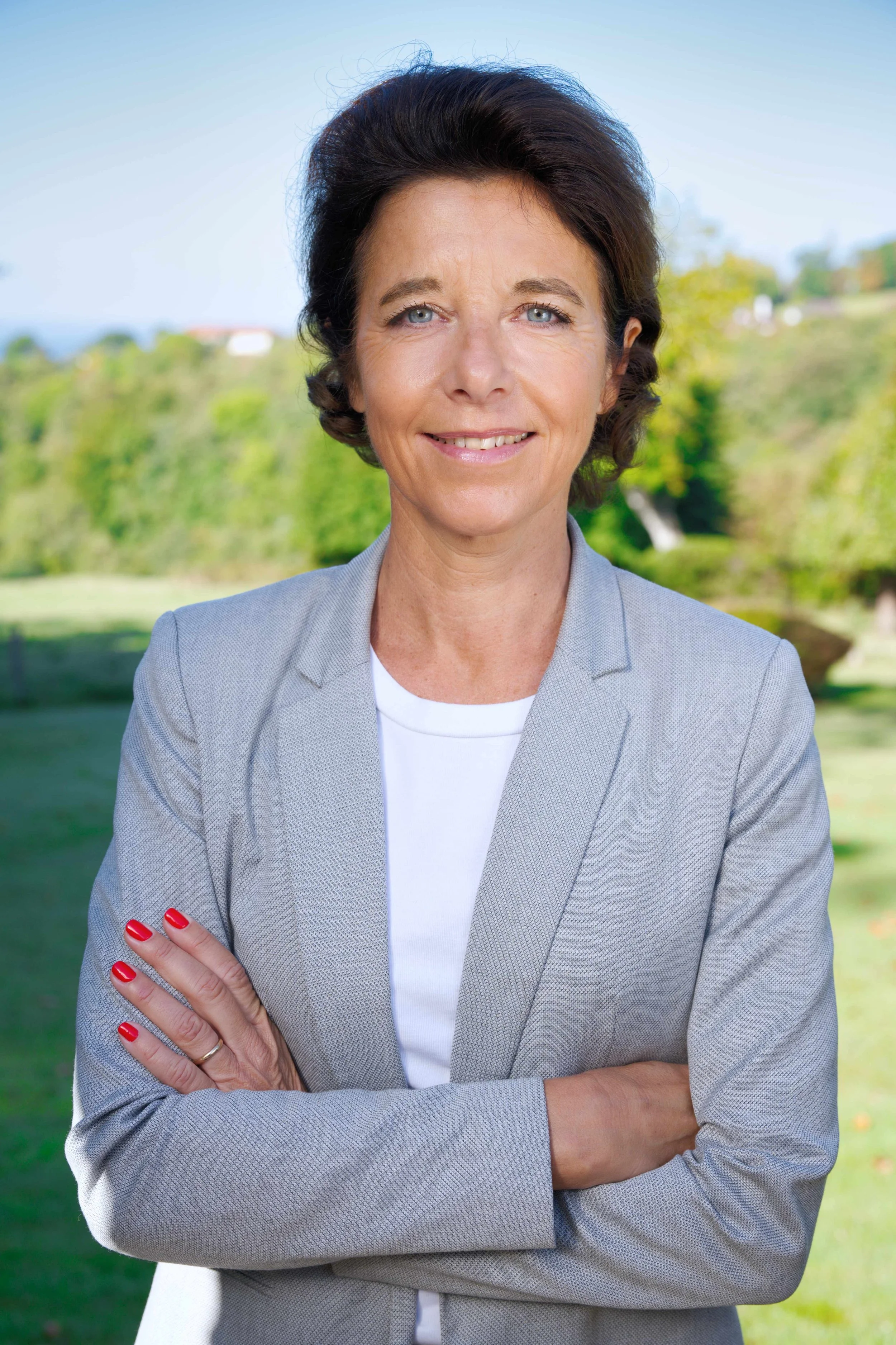 Femme souriante en chemisier gris et veste grise, bras croisés, dans un parc avec arbres verts en arrière-plan.
