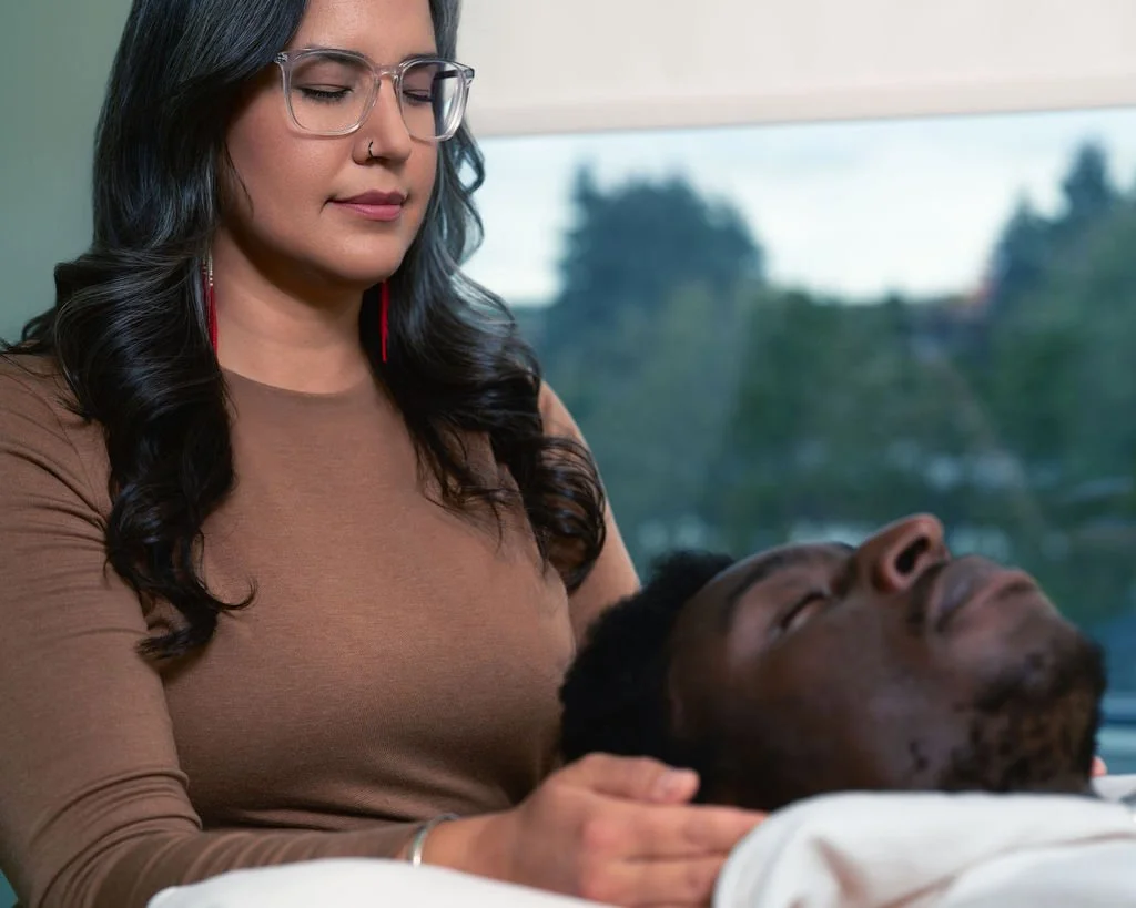 Woman with glasses and dark hair caring for a man with closed eyes, lying in a hospital bed.