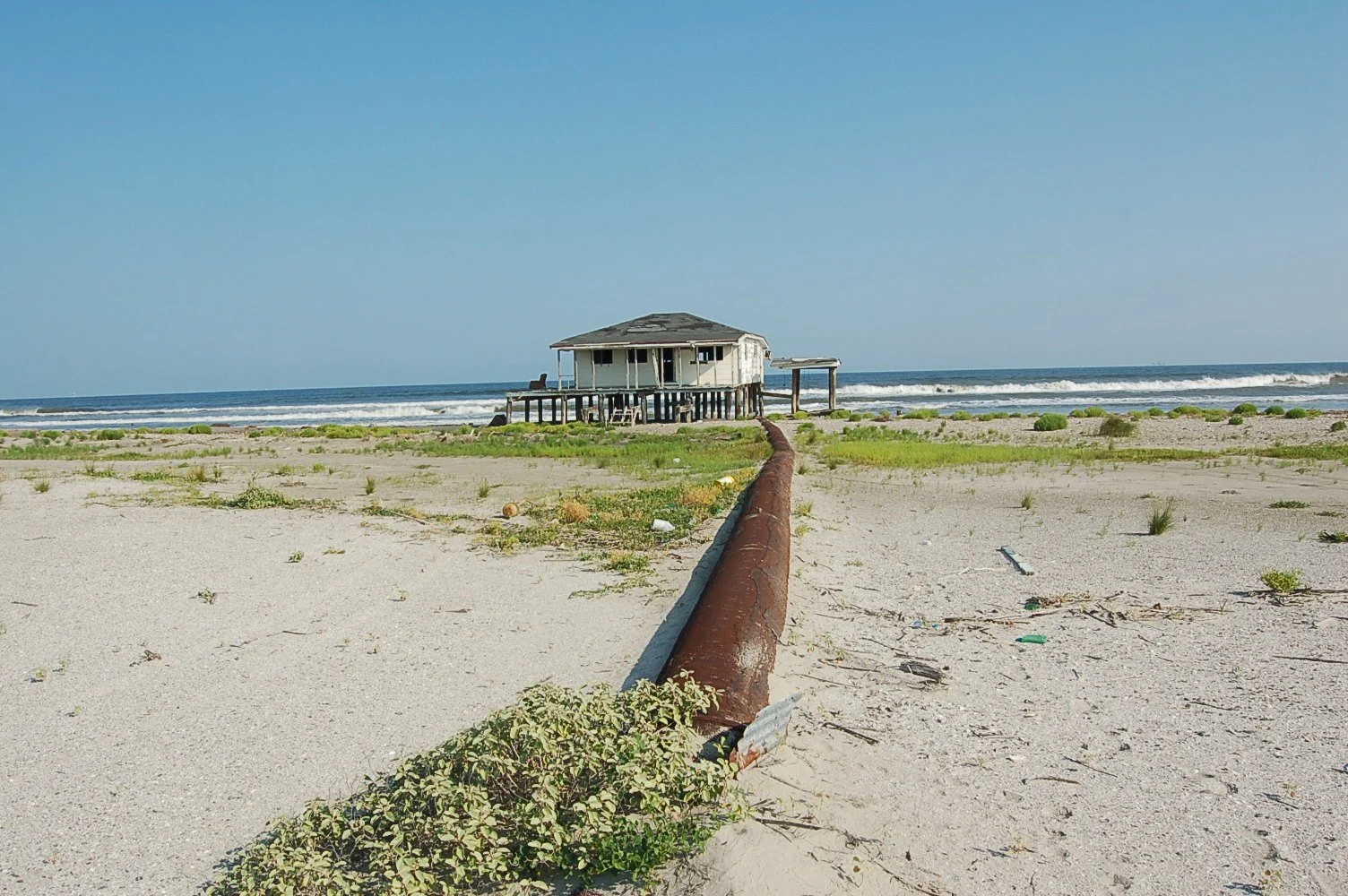 An image of a lifted house on a sandy coast with the ocean in the distance under a clear blue sky.