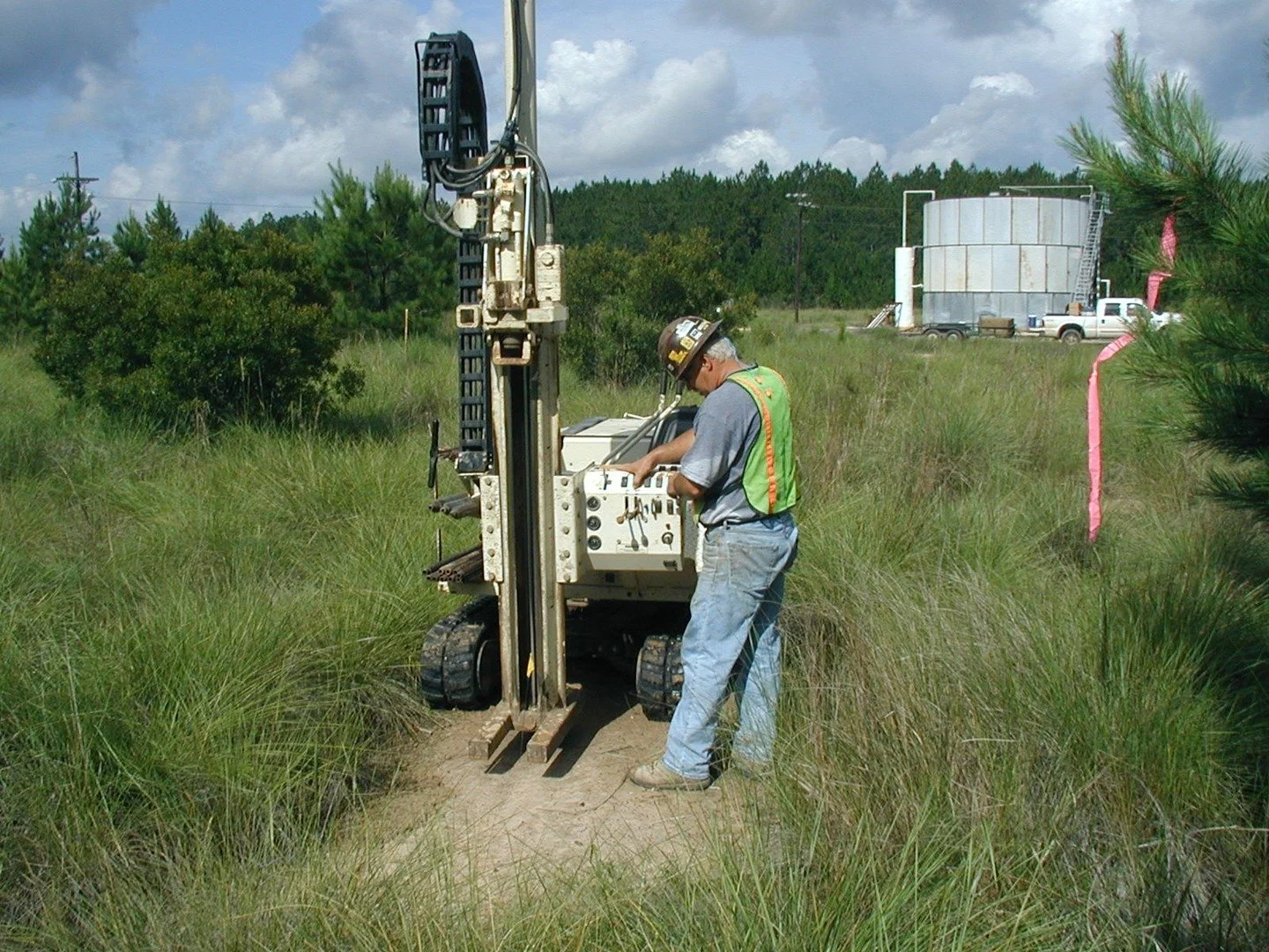 A man wearing personal protective equipment and operating a machine in a grassy field with green foliage behind him under a cloudy sky.
