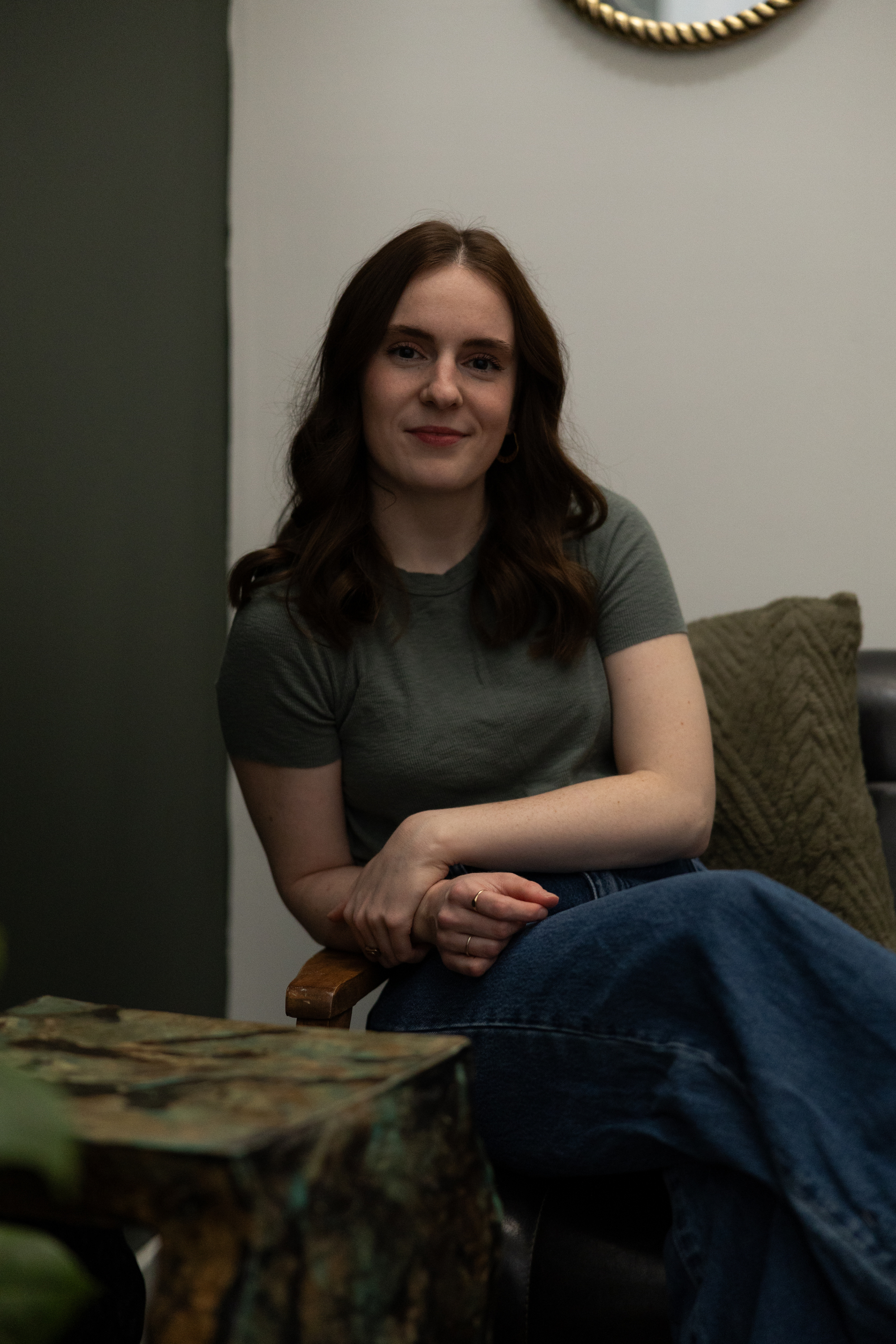 A woman with shoulder-length brown hair, wearing a gray t-shirt and blue jeans, sitting on a black chair with her arms crossed, smiling at the camera in a cozy room.