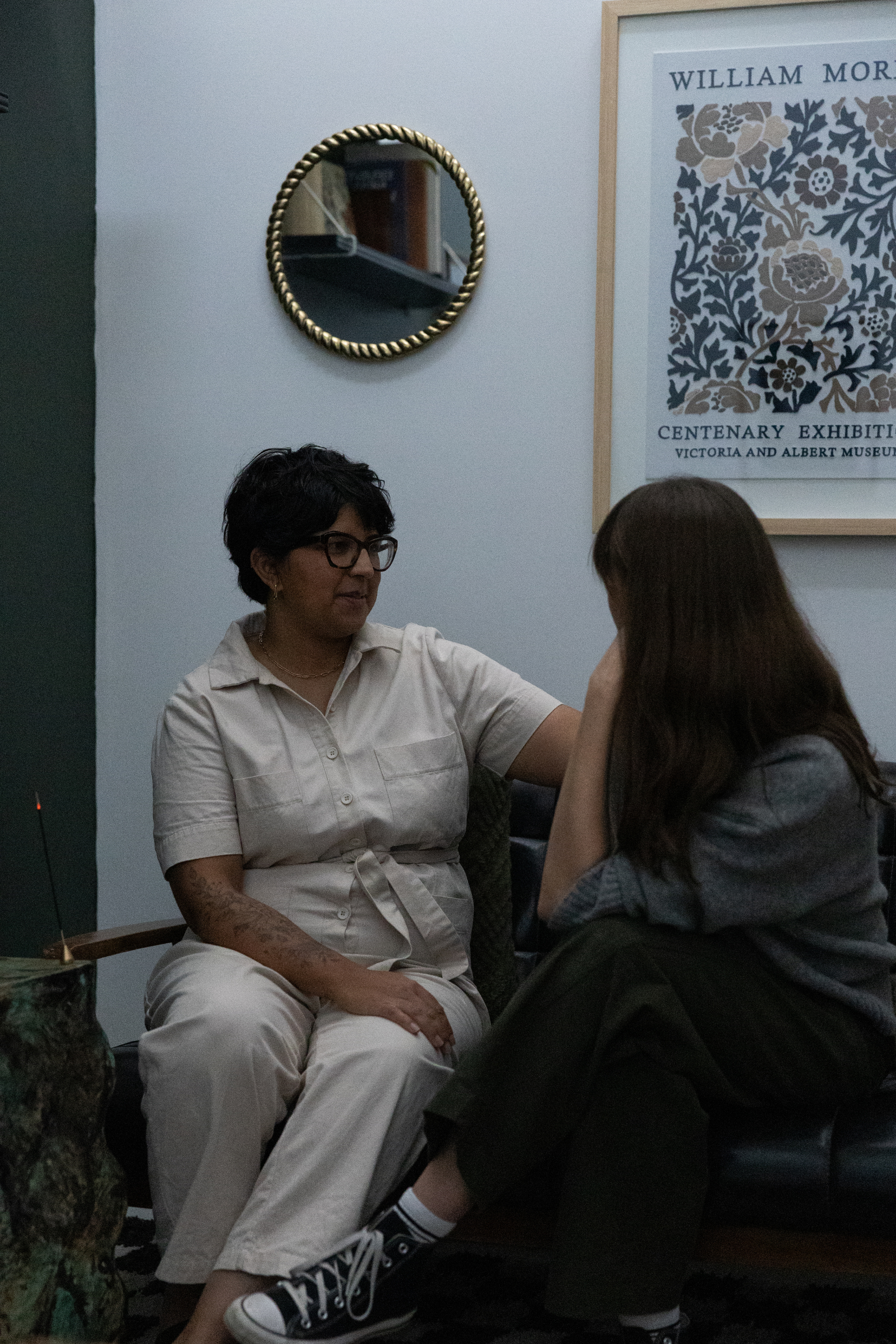 Ayisha, a Calgary-based occupational therapist with short black hair, glasses, and tattoos on her arms, comfortingly touches a young girl who is sitting with her head bowed and her hand covering her face, in a room with framed art and a mirror.