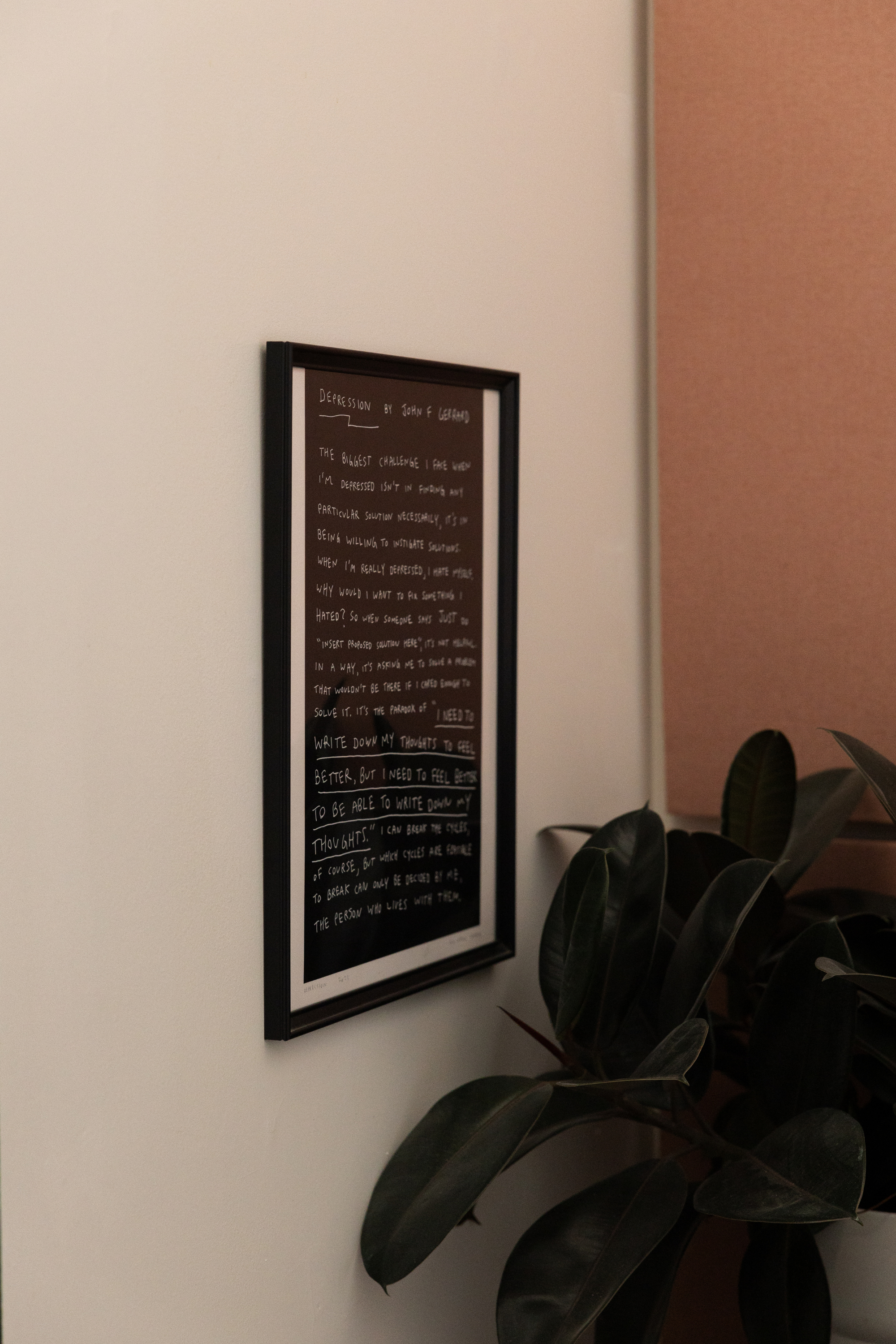 A black framed chalkboard-style poster or sign with white handwritten text, mounted on a white wall next to a beige wall with a leafy green plant in front.