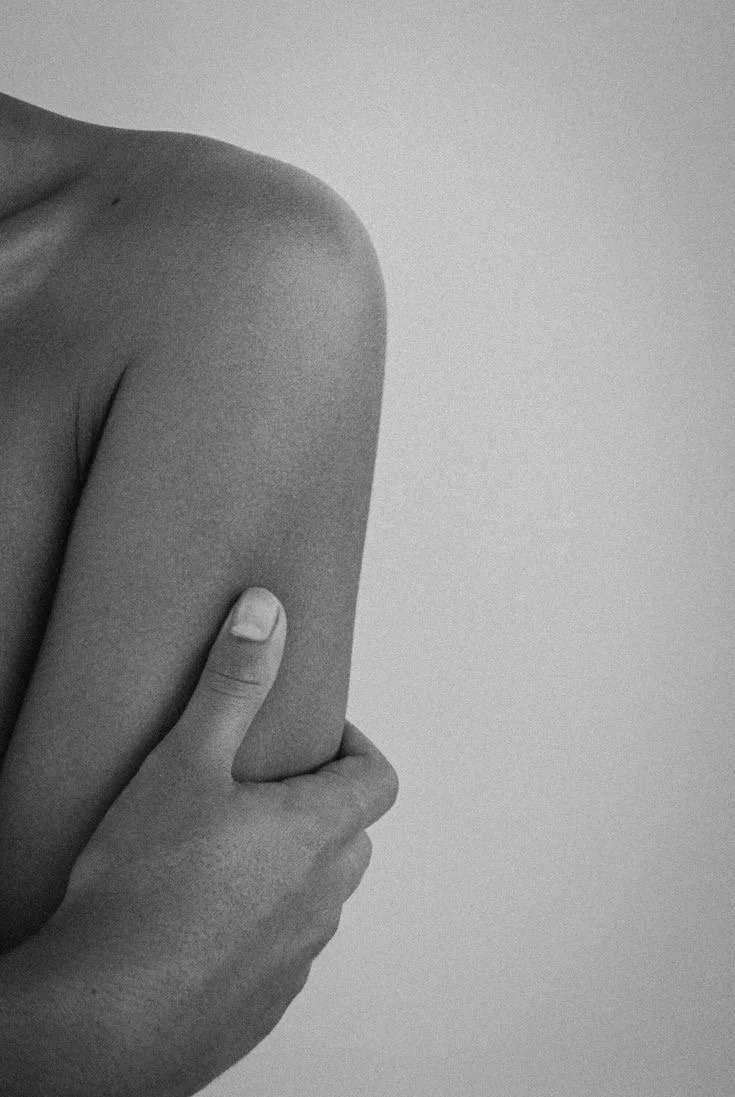 A black-and-white close-up photo of a person's shoulder and arm, with their hand gently holding their upper arm.