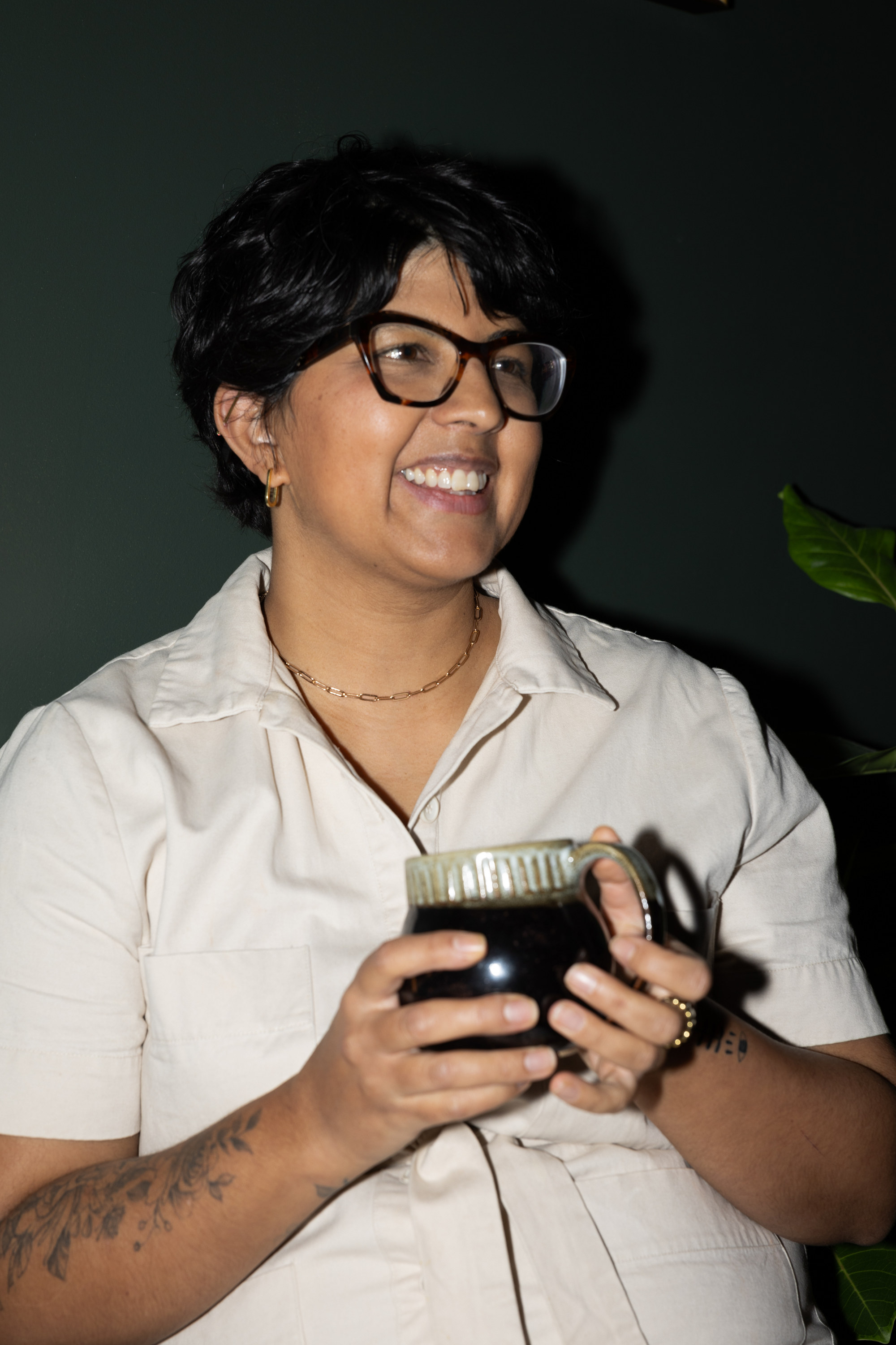 A woman with short black hair, glasses, and earrings, smiling while holding a dark mug with a beige top.