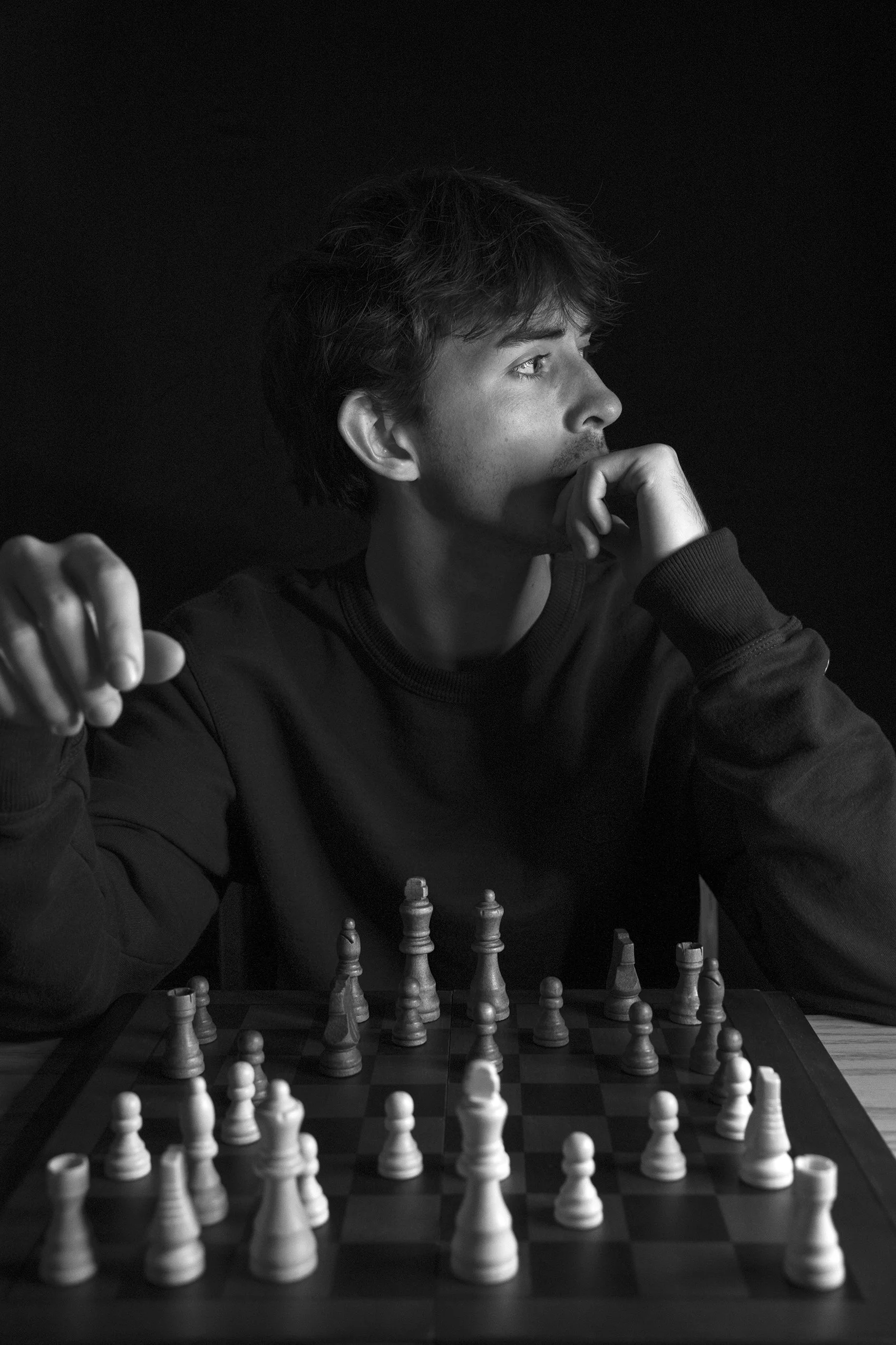 Black and white photo of a young man with tousled hair, looking thoughtfully to the side while playing chess. His hand is up to his chin, and the chessboard with pieces is in front of him against a dark background.