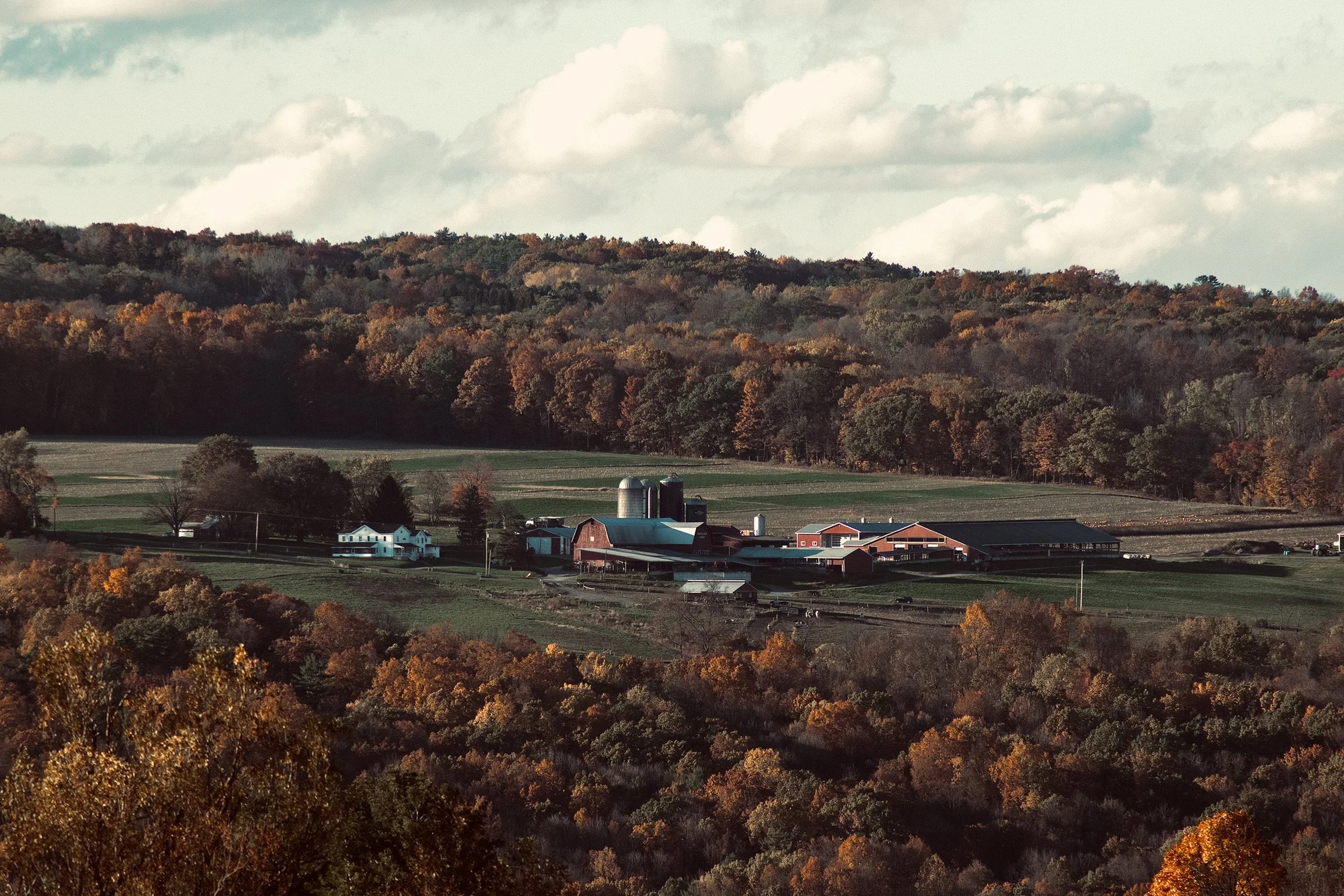 A rural farm with red barn buildings, silos, a white house, and fields surrounded by trees with fall foliage in the mountains.
