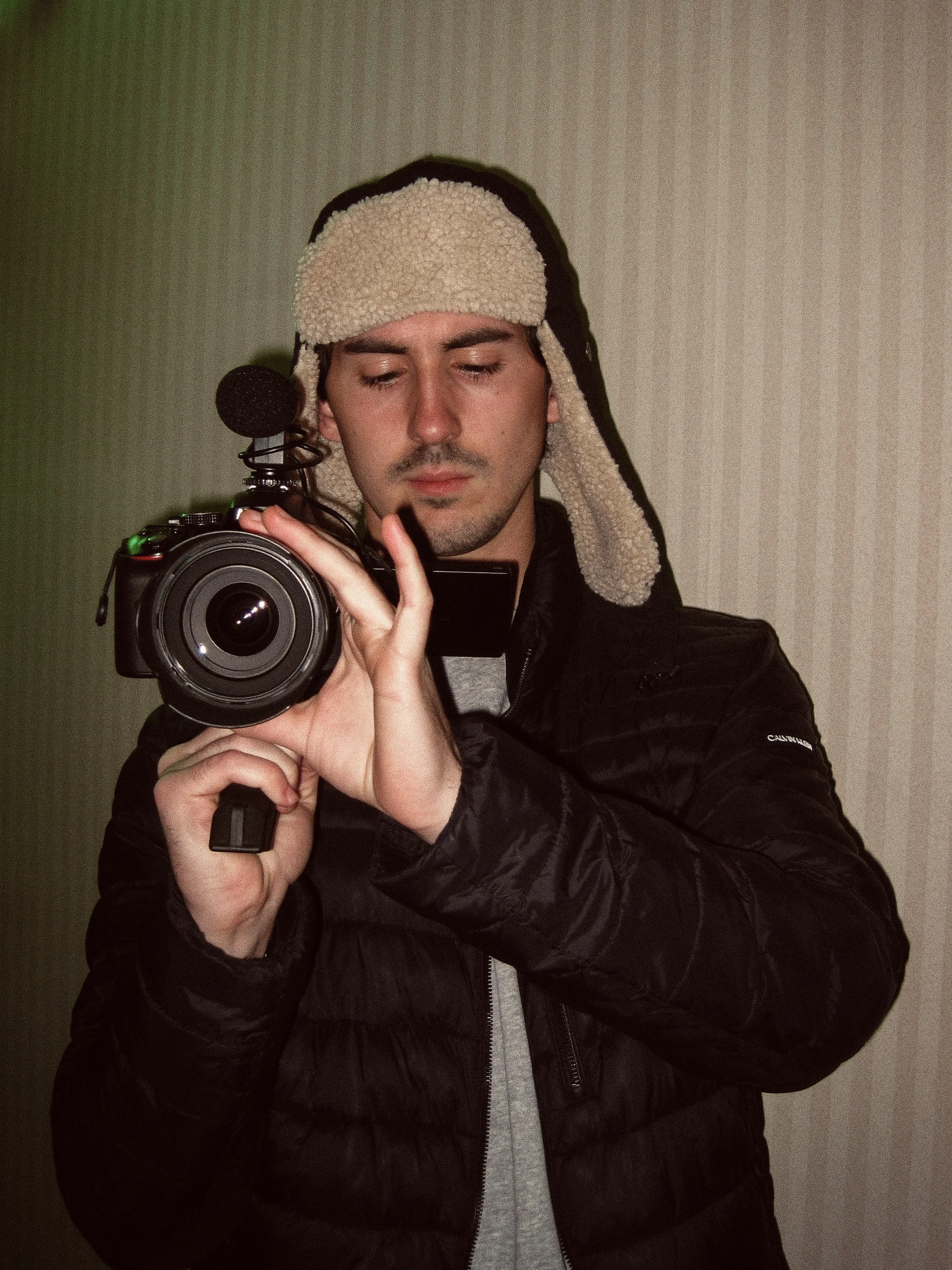 A man taking a selfie with a camera, wearing a warm hat with ear flaps and a black puffy jacket.