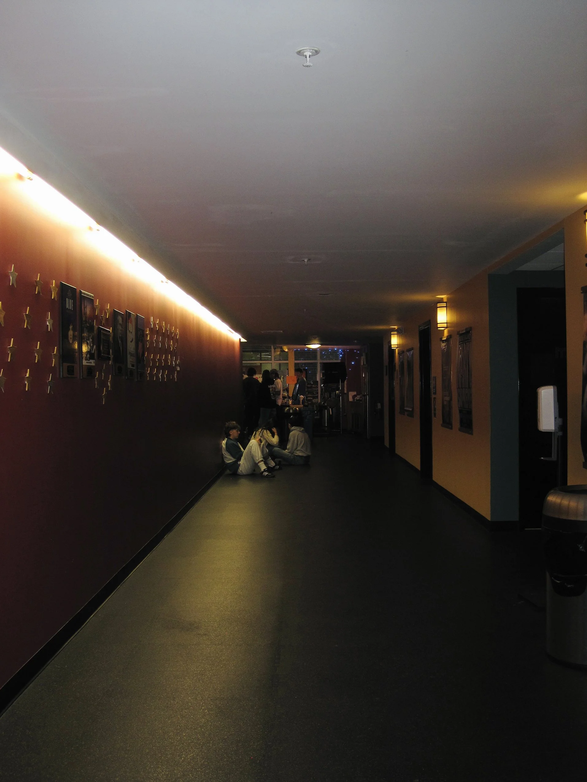 Dimly lit indoor hallway with children sitting on the floor near the wall, some people standing and talking in the background, and movie posters on the walls.