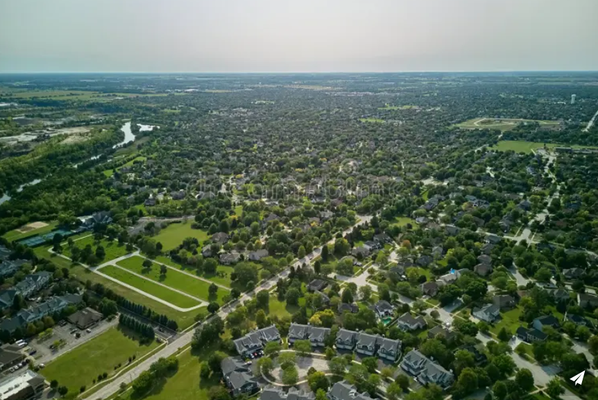 Vue d'ensemble d'une banlieue résidentielle avec de nombreuses maisons, arbres et routes, au bord d'une rivière, sous un ciel clair.
