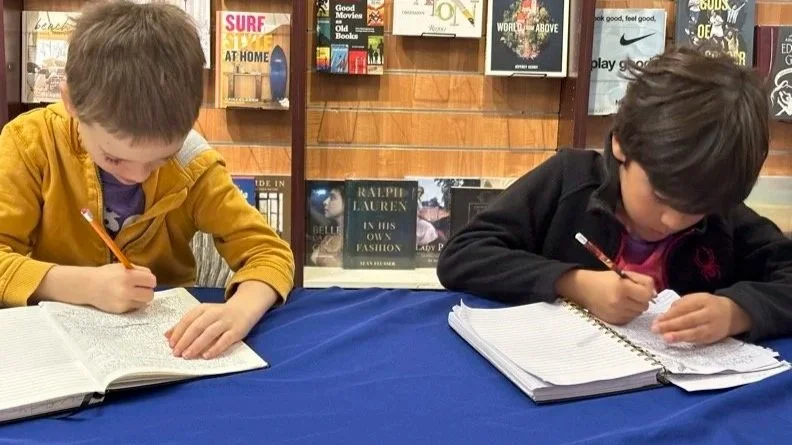 Writewithconfidence - Middle schoolers sitting at a table in a library, reading and writing in notebooks with pencils.