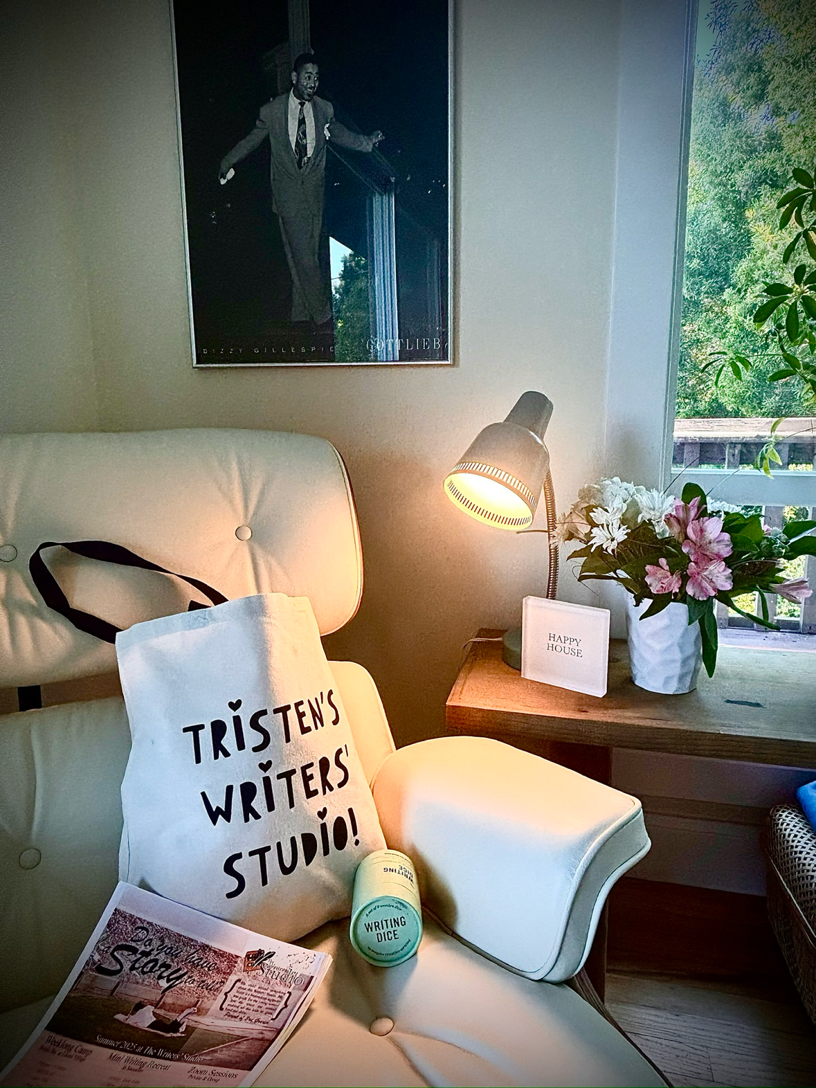 'TRISTEN'S WRITERS STUDIO!', a magazine, and a cylindrical container labeled 'WRITING DICE'. A wooden side table with a lamp, a framed note reading 'HAPPY HOUSE', and a white vase with pink and white flowers sit beside a window with green foliage outside. A black-and-white poster of a man in a suit is on the wall above.
