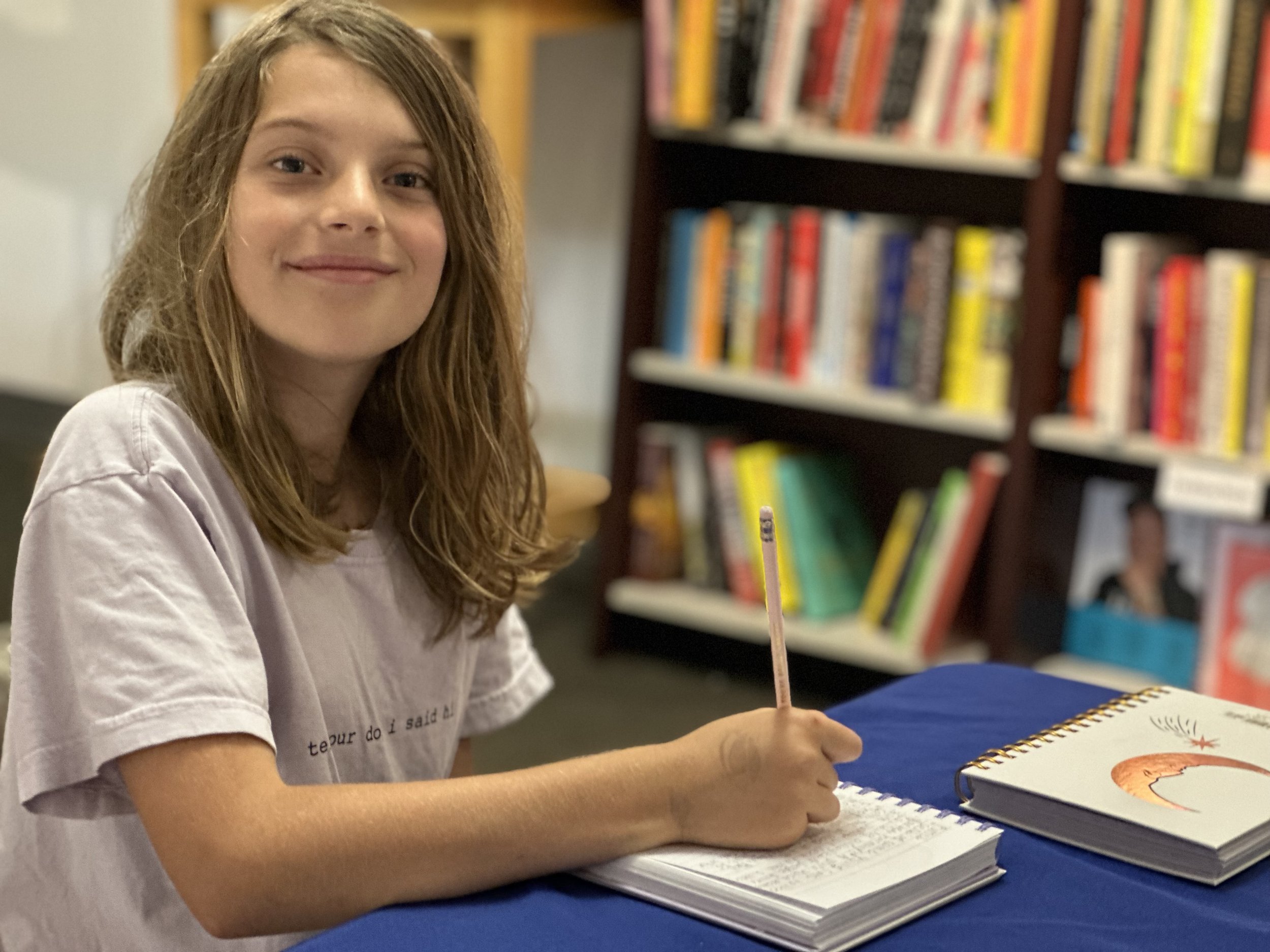 A writer ready to work in an open notebook. There is a spiral-bound sketchbook with a drawing of a crescent moon and a star on the cover, placed on the table. In the background, there is a bookshelf filled with various colorful books.