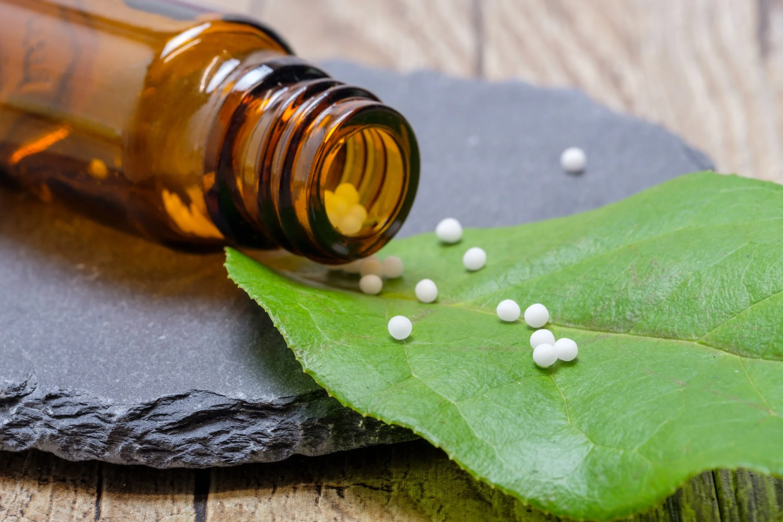 An amber glass bottle tipped over on a green leaf, with small white homeopathy pellets spilling out onto the leaf and a dark stone surface.