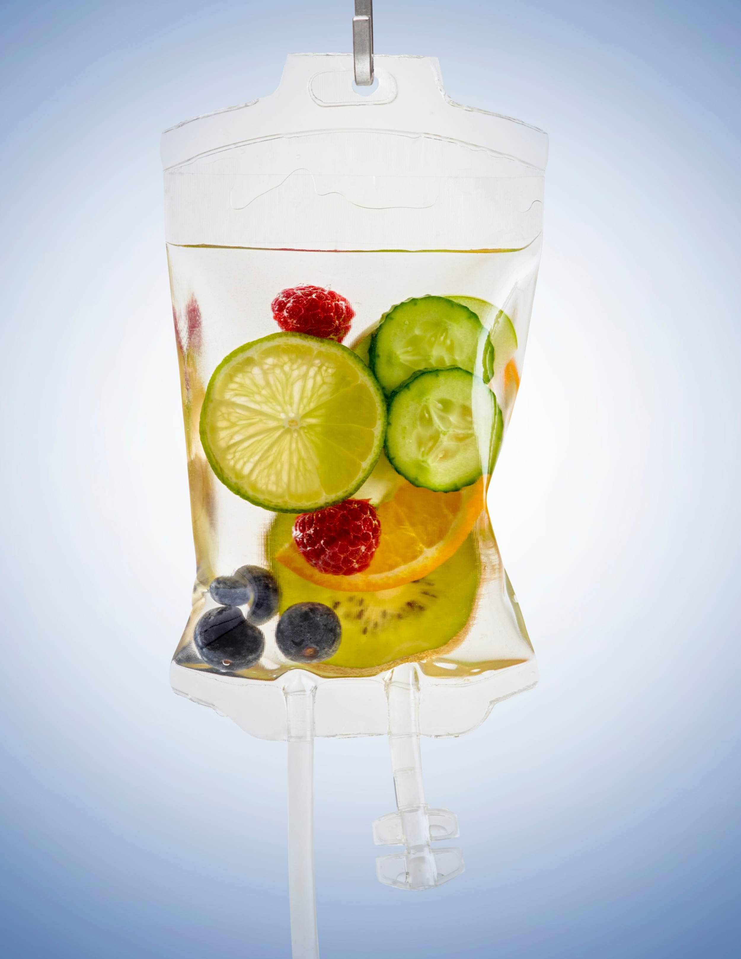 A medical IV drip filled with slices of lemon, cucumber, raspberry, blueberries, and orange in clear liquid.