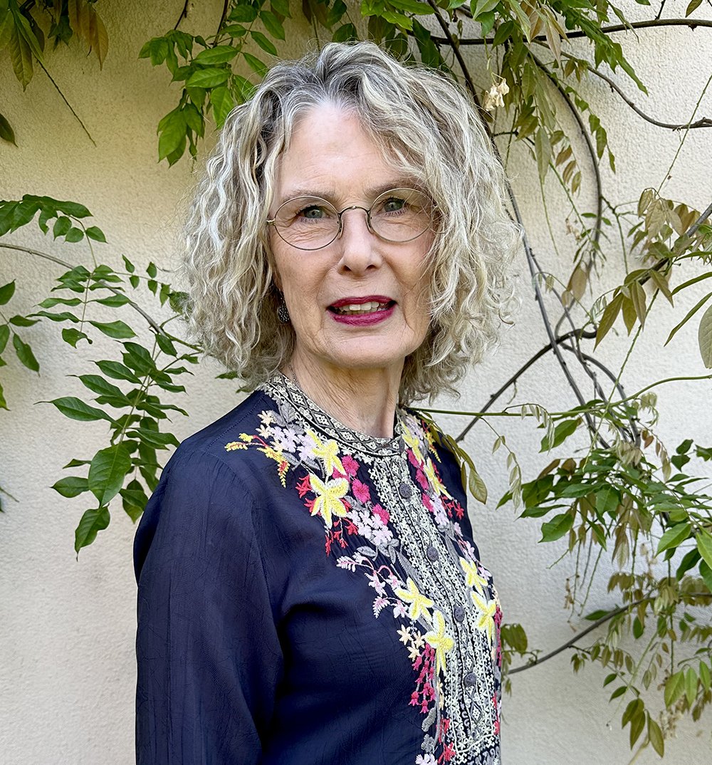 A middle-aged woman with curly gray hair and glasses, wearing a navy blouse with colorful floral embroidery, standing outdoors in front of a wall with green leafy vines.