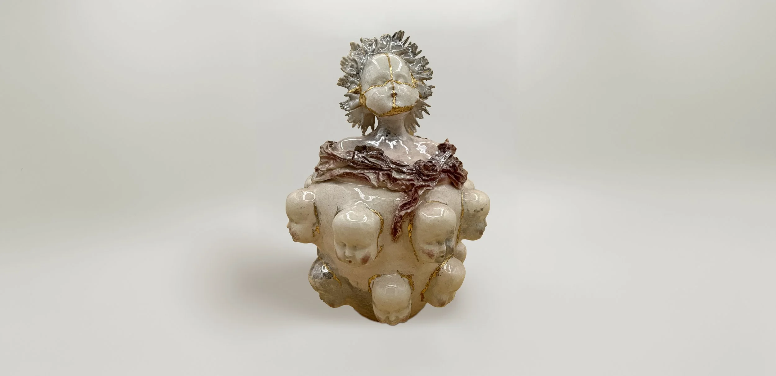 Ceramic sculpture of a figure with a face mask, surrounded by multiple small faces, featuring metallic accents and a flowing cloth, set against a plain white background.