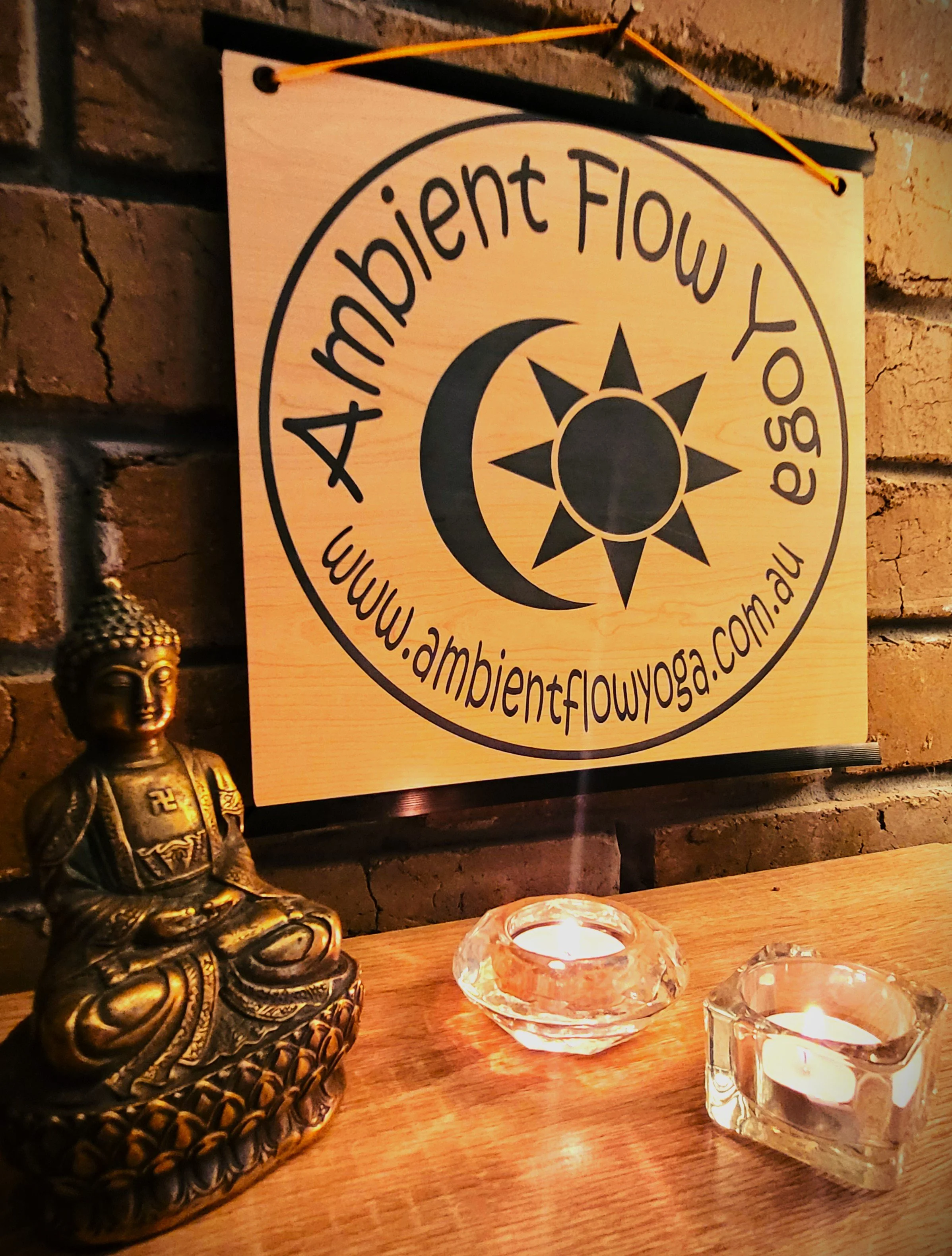 A wooden sign with the text 'Ambient Flow Yoga' and the website 'www.ambientflowyoga.com.au' inside a circle, featuring a crescent moon and sun icon. On a wooden surface in front of the sign, there is a small Buddha statue, a round glass candle holde