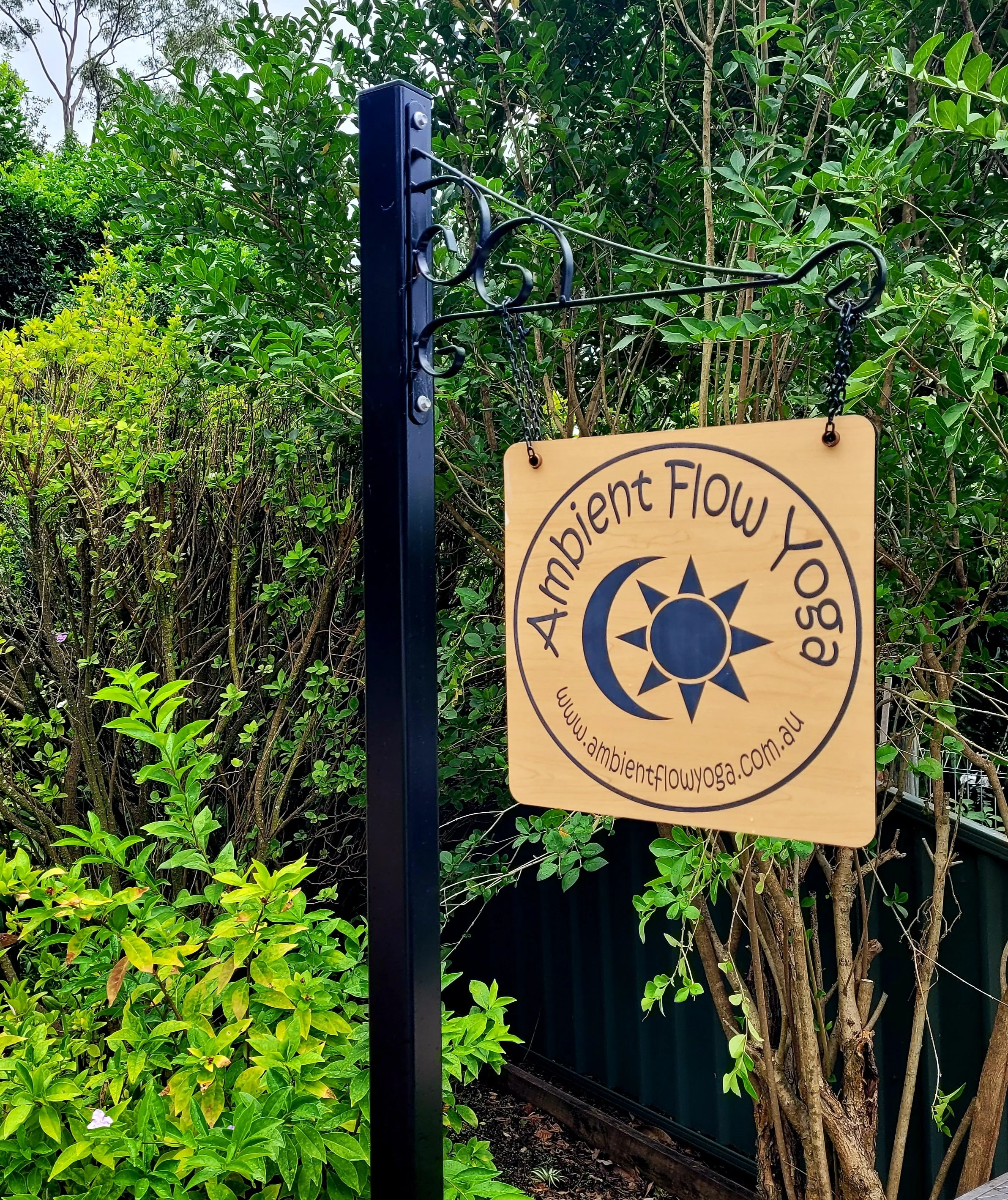Wooden sign hanging from a black metal stand that reads "Ambient Flow Yoga" with a website URL, surrounded by green bushes and trees.