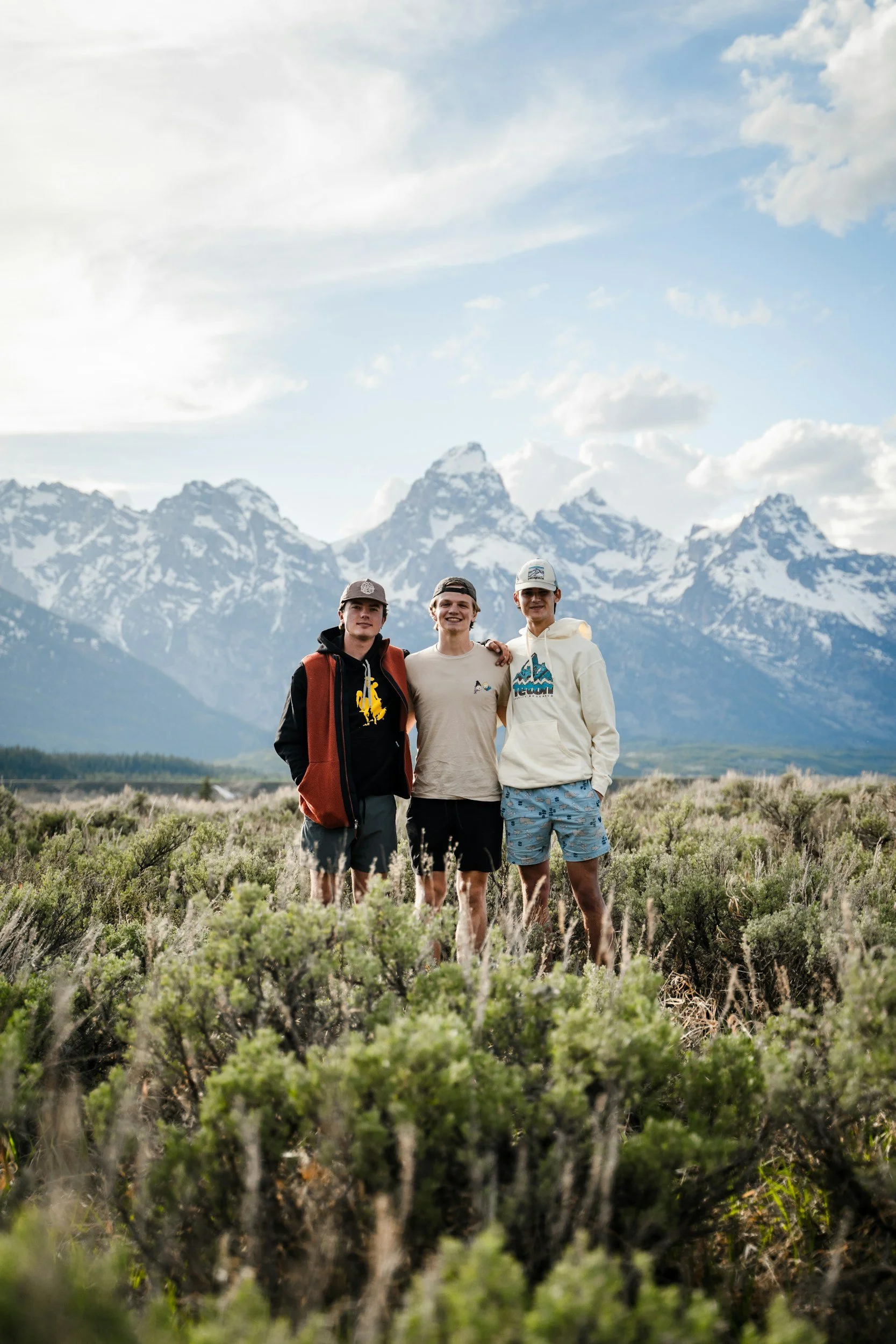 Three young men standing together outdoors in a field with snow-capped mountains in the background.