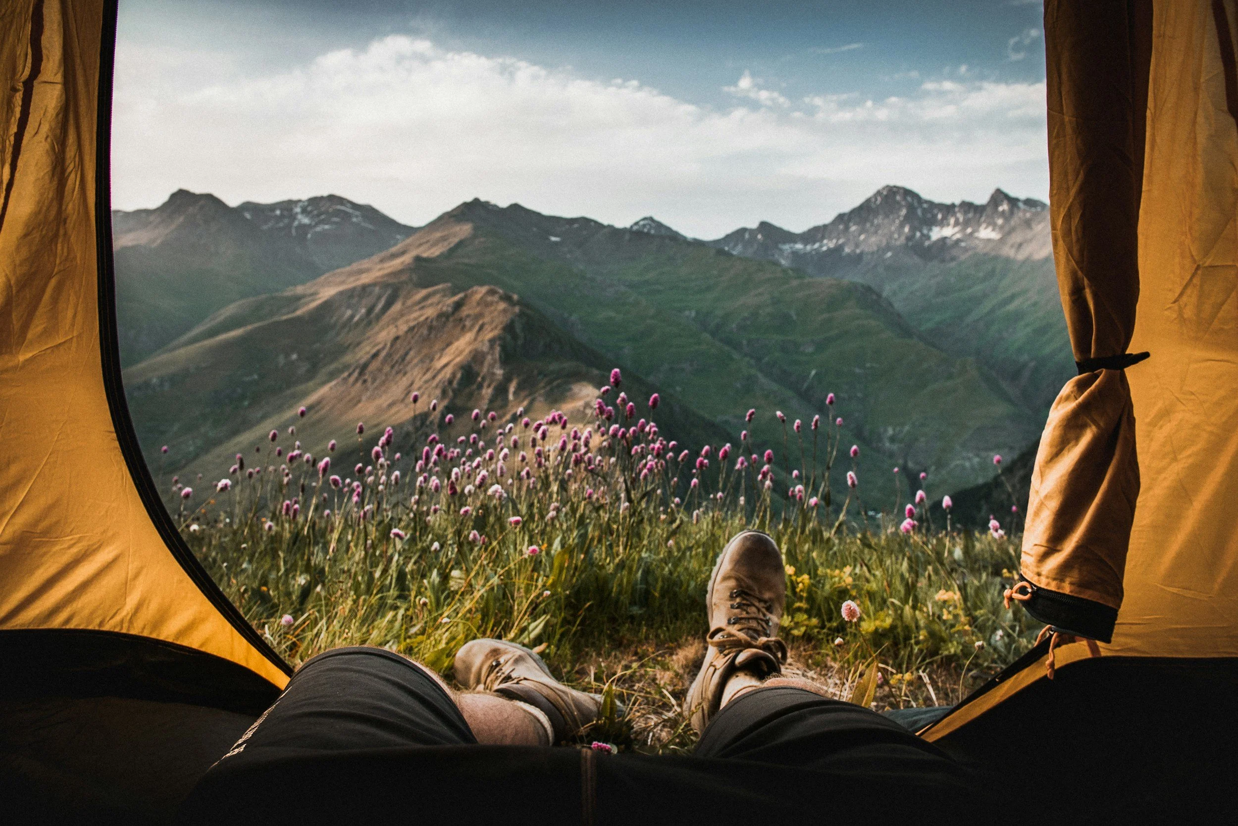 View from inside a yellow camping tent showing a person relaxing with legs stretched out, overlooking a scenic mountain landscape with green hills and pink wildflowers.