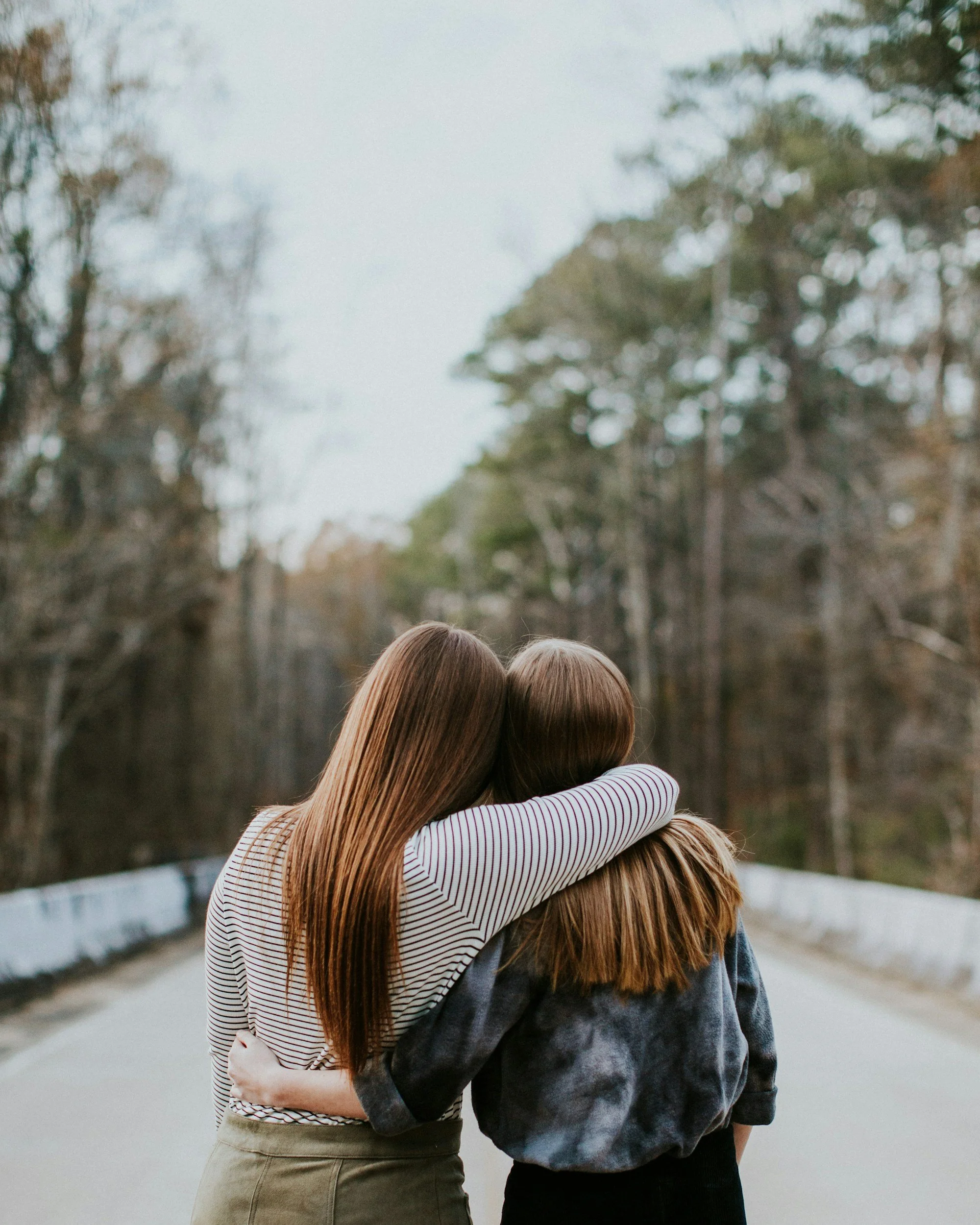 Two women hugging on a road surrounded by trees.