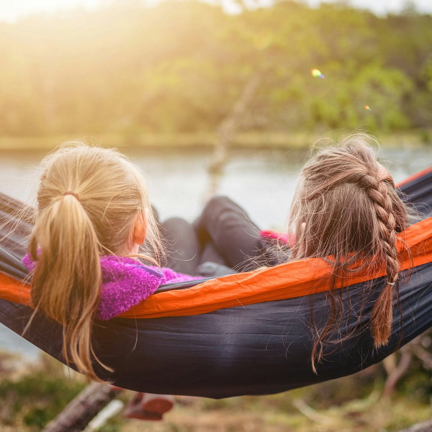 Two girls sit in a hammock by a lake.